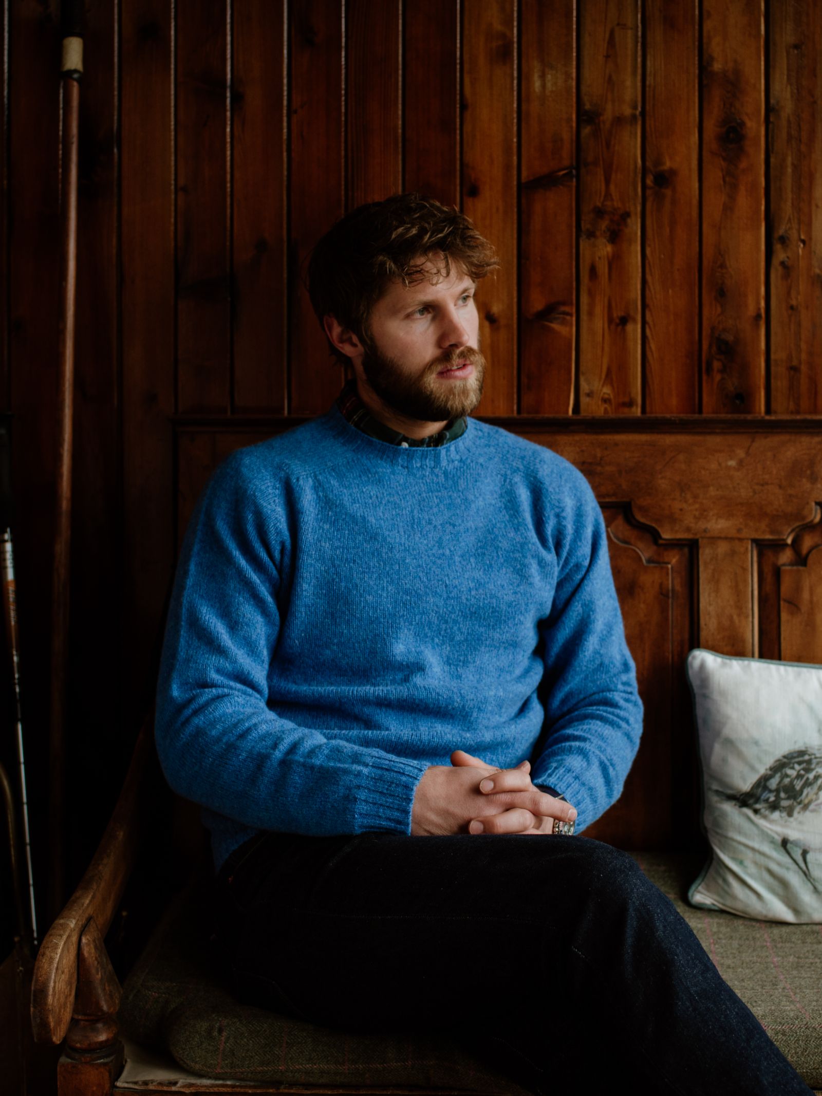 A bearded man with brown hair sits on a wooden bench against wood paneling, wearing a blue Shetland Jumper by Campbells of Beauly, crafted from soft lambswool yarn in heritage-inspired colourways, hands clasped as he gazes thoughtfully aside.