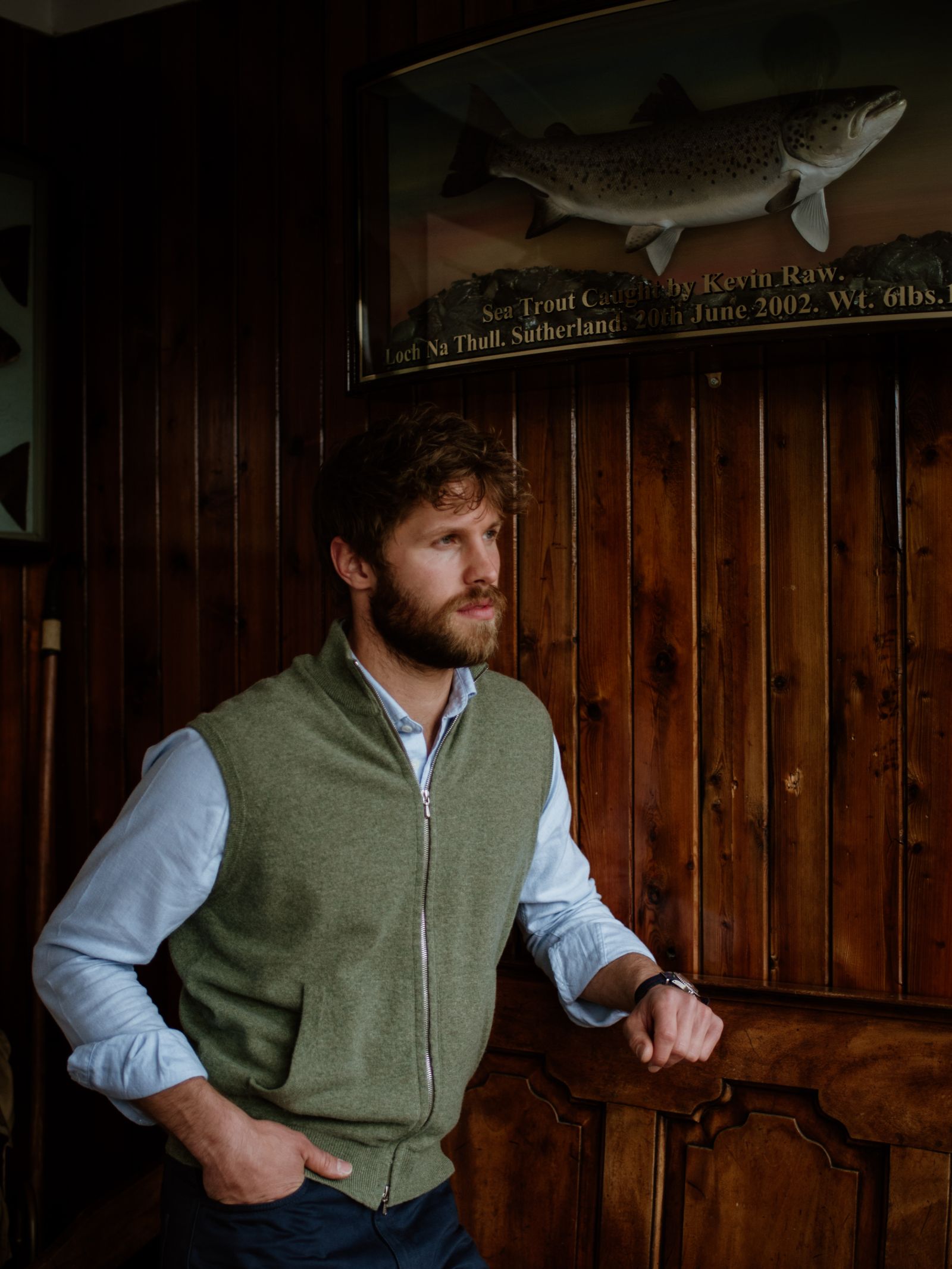 A man with a beard and light brown hair stands indoors against a wooden wall, wearing a light blue shirt and the Campbells of Beauly Zip Gilet. Above him, a mounted fish display with an inscription enhances the timeless setting.