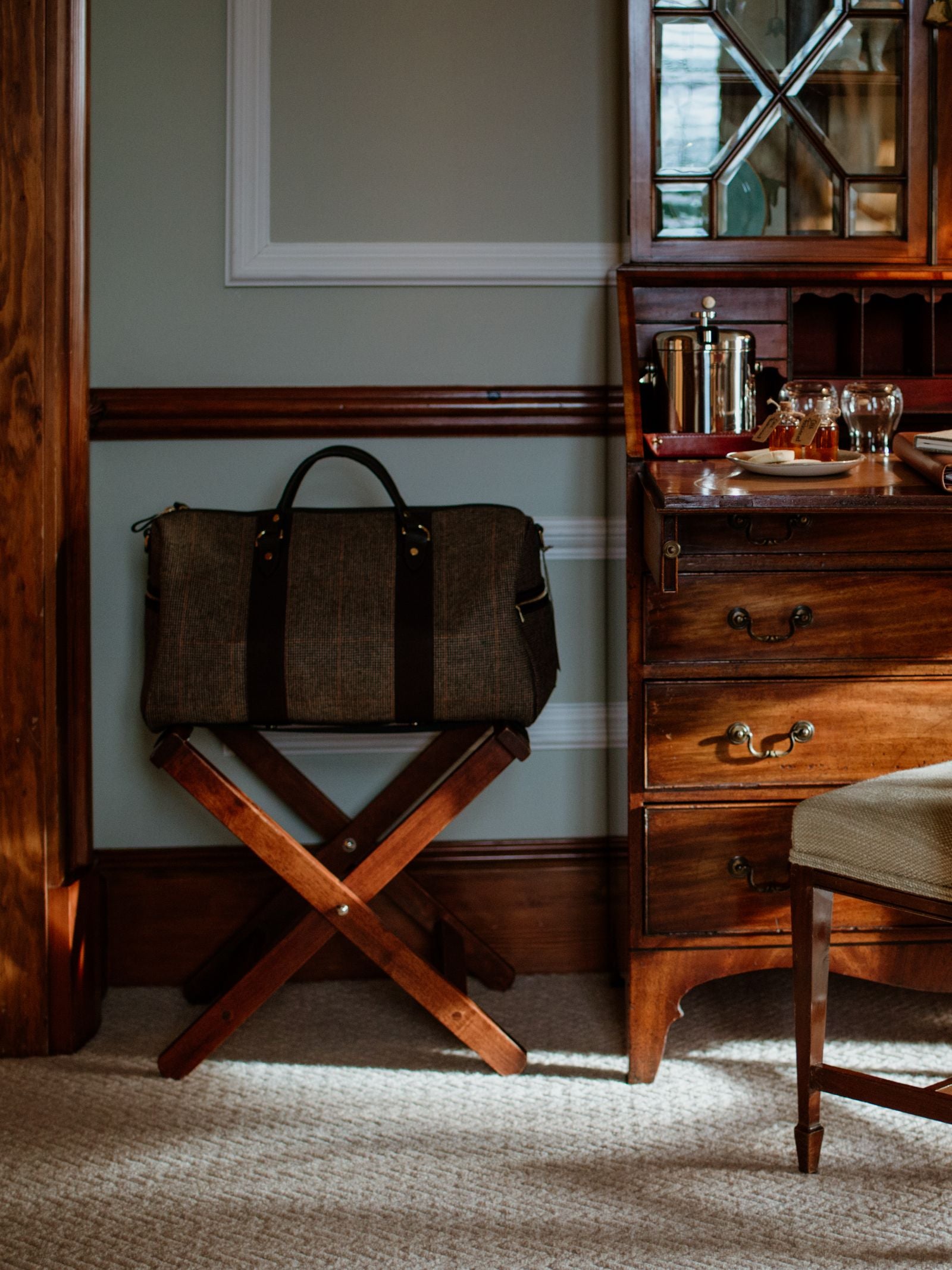 A Campbell's of Beauly Hand Luggage Holdall in brown stripes rests on a wooden rack beside a classic desk with drinks and glasses—the ideal travel companion for any journey.