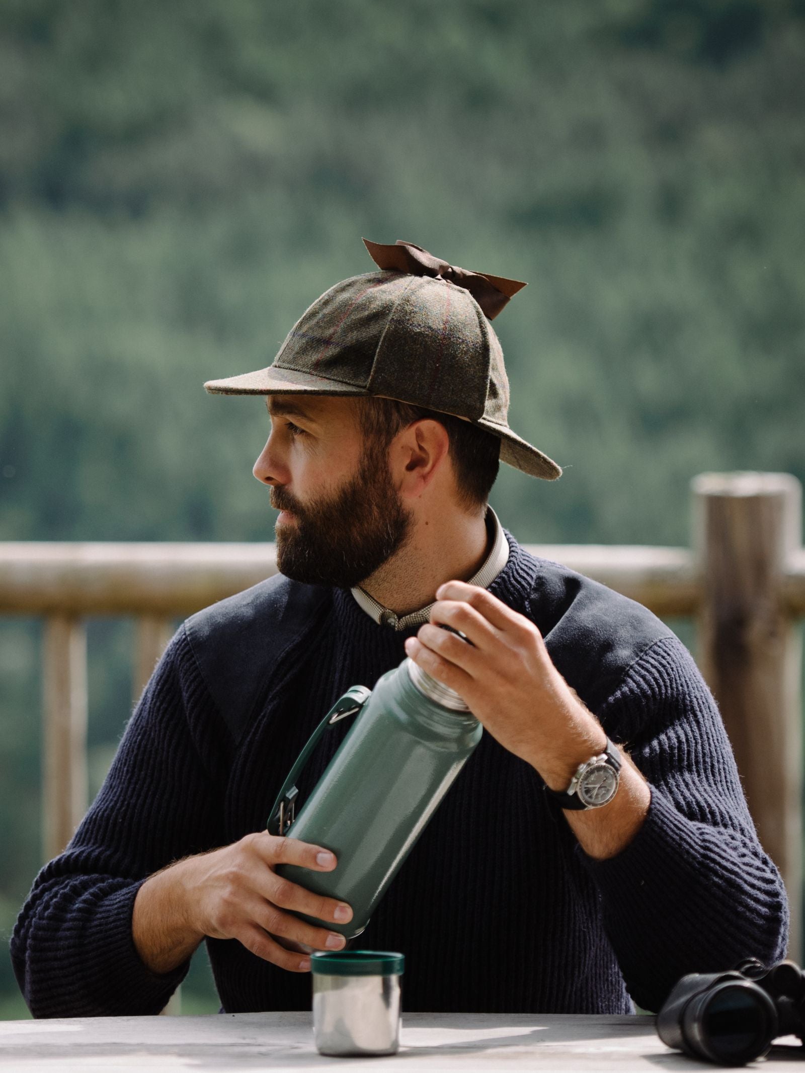 A bearded man in a hat and Campbells of Beauly Scotia Suede Jumper sits at an outdoor table with a green thermos, cup, and binoculars. A blurred forest forms the background.