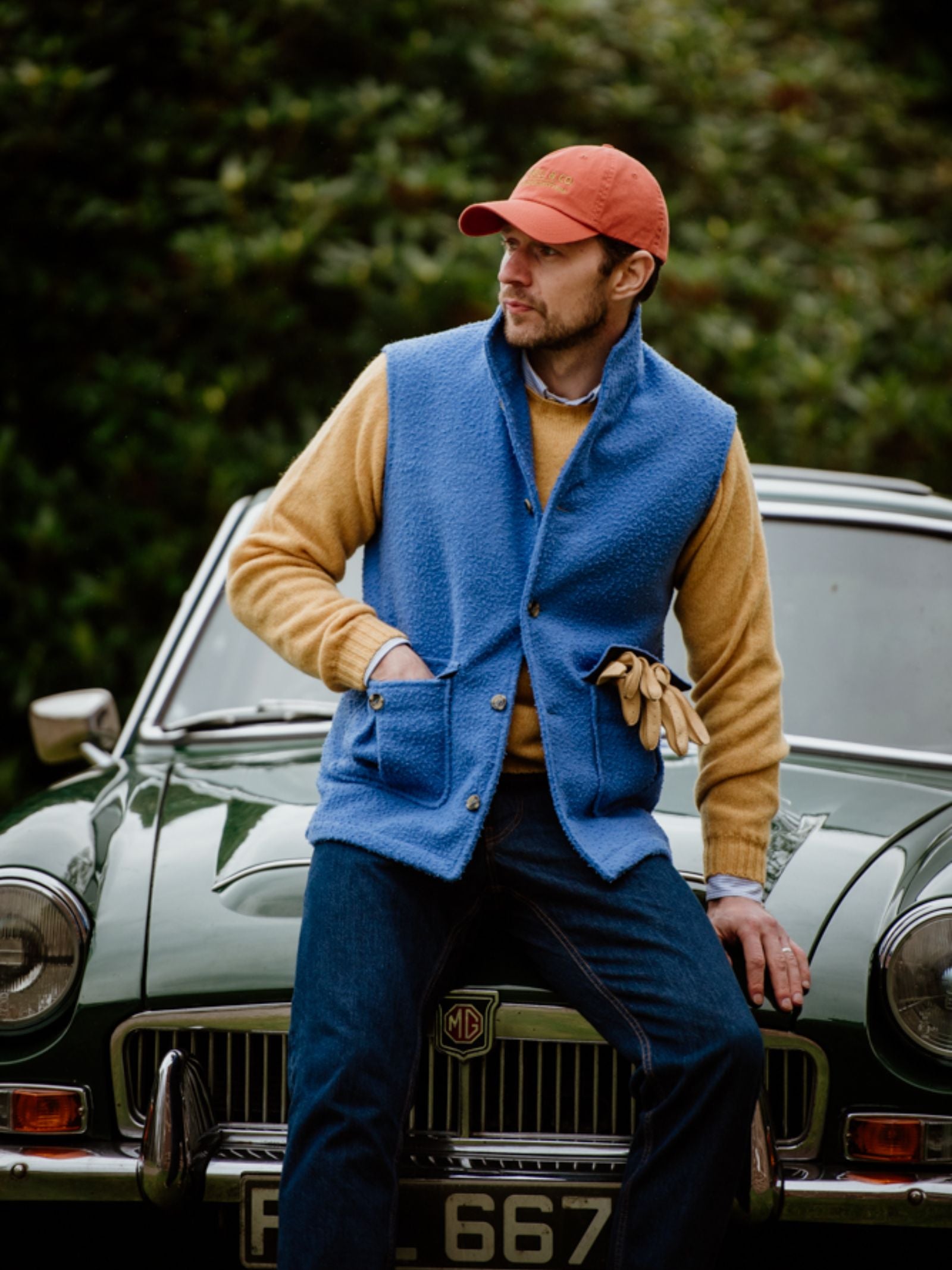 A man in a red cap, yellow sweater, and Campbells of Beauly Casetino Fleece Gilet sits on the hood of a classic green MG car, holding gloves, with greenery in the background.