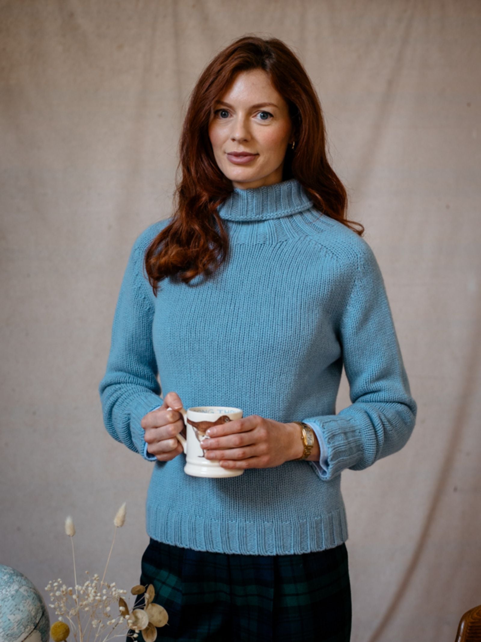 A woman with long auburn hair wears the Campbells of Beauly Chunky Roll Neck Sweater in light blue, holding a mug with both hands, standing before a beige backdrop with dried flowers in the lower left corner.