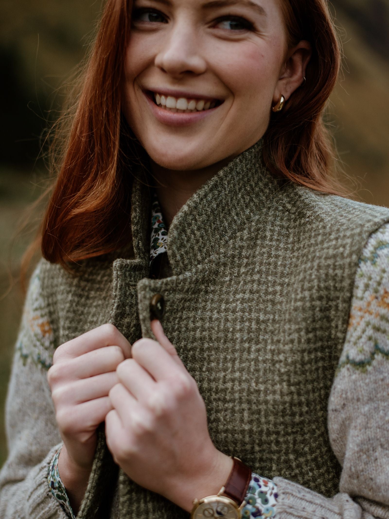 A woman with long red hair smiles while holding the collar of her Campbells of Beauly Harris Tweed Houndstooth Gilet, displaying quality tailoring. She pairs it with a floral shirt, gold hoop earrings, and a brown leather watch outdoors.