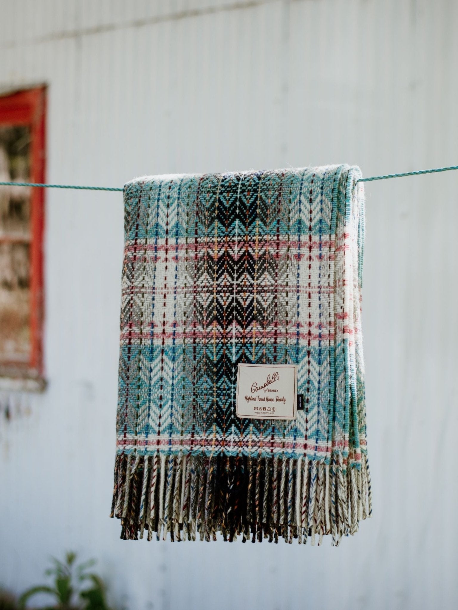 The Wyvis Lambswool Throw by Campbells of Beauly, featuring blue, brown, and red stripes with fringe, is draped over an outdoor clothesline against a blurred white wall and red-trimmed window.