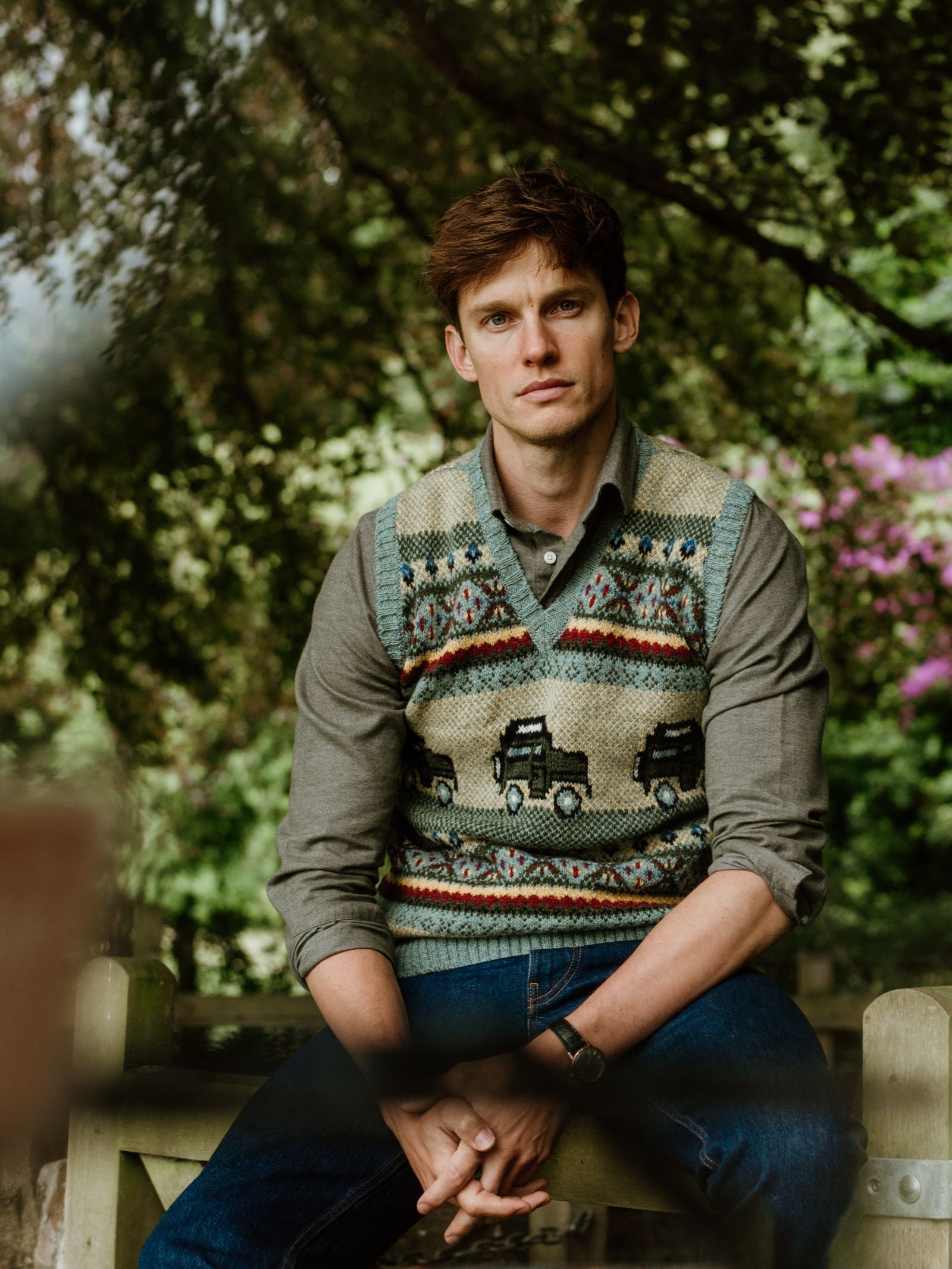 A young man with short brown hair sits on a wooden bench outdoors, wearing the Land Rover Defender Fairisle Slipover by Campbell’s of Beauly over a gray shirt, surrounded by lush greenery as he gazes thoughtfully at the camera.
