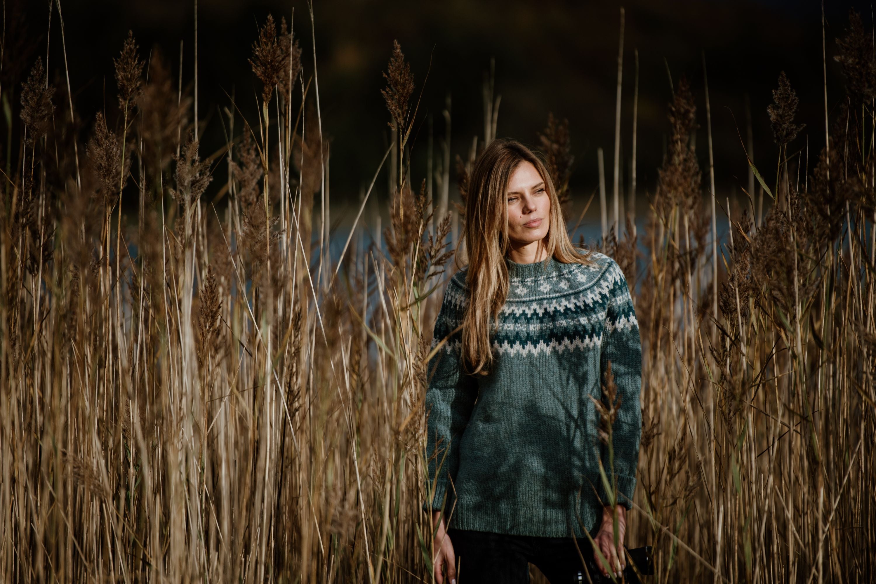 A woman with long hair wearing a patterned sweater stands among tall, dry reeds outdoors, with sunlight casting shadows on her.