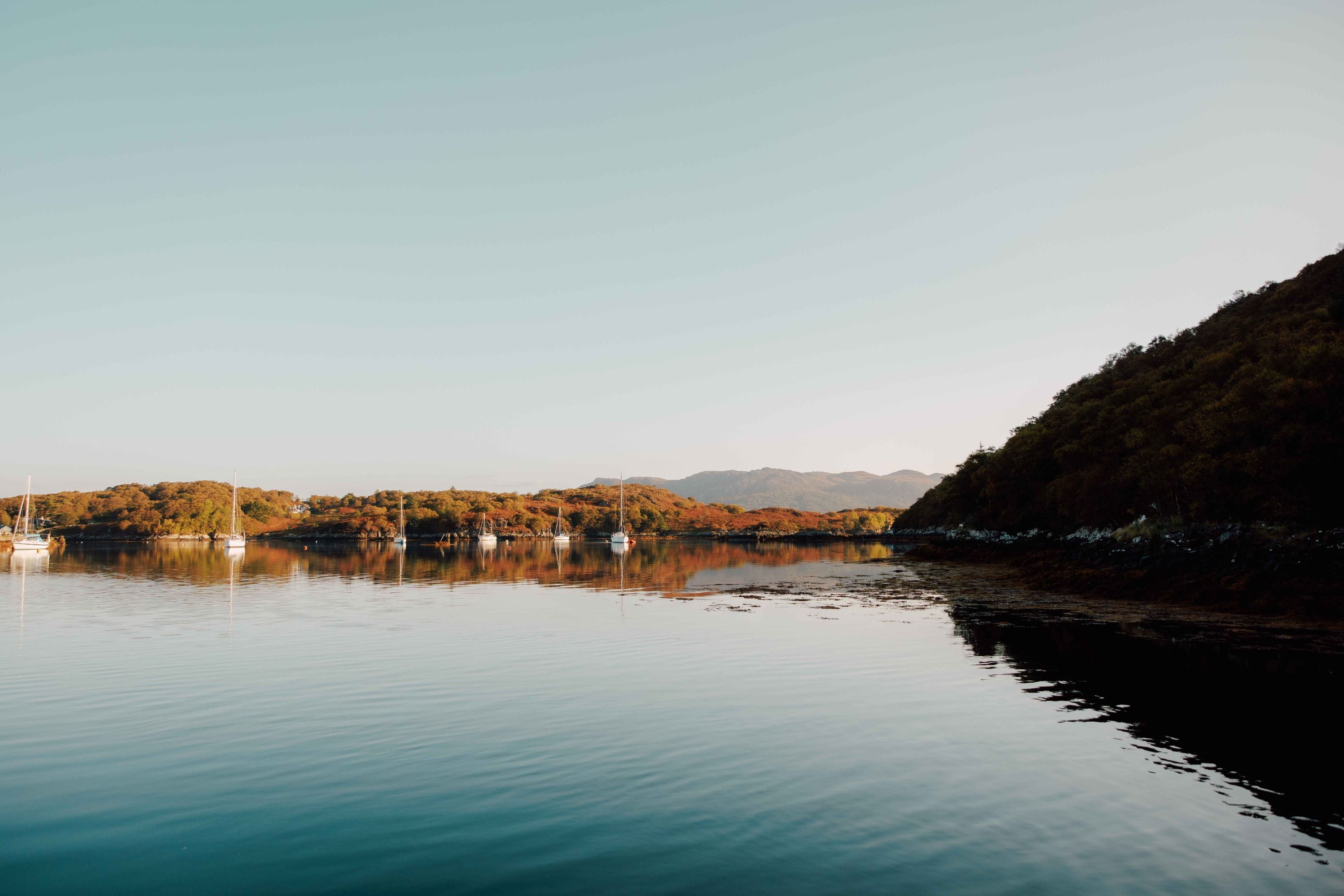 A calm lake reflects the surrounding hills and trees under a clear sky, with several small boats anchored near the shore. The scene is peaceful and bathed in soft, natural light.