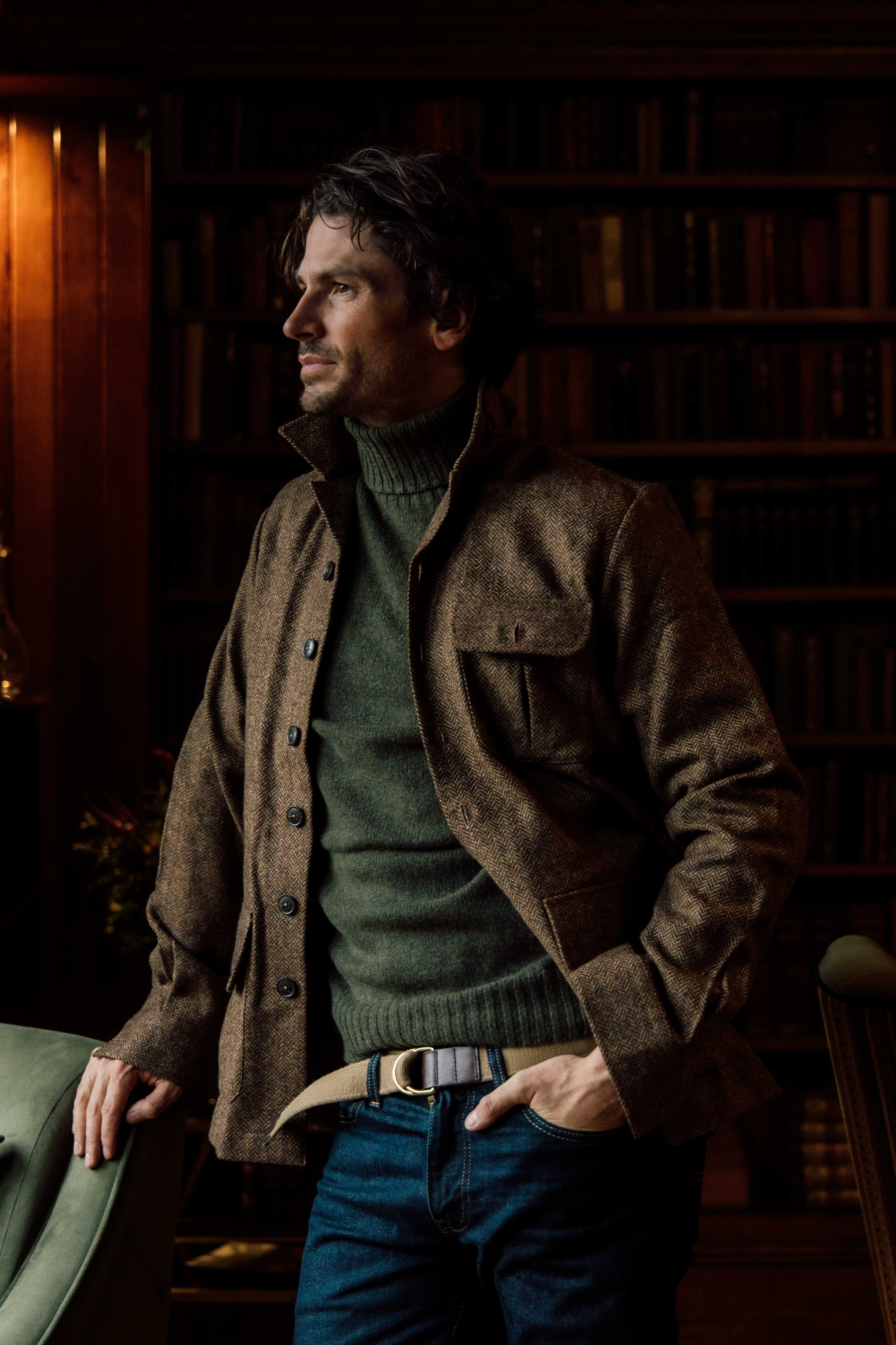 A man with wavy dark hair wearing a brown jacket over a green turtleneck sweater stands indoors, leaning on a green armchair, with shelves of books blurred in the background.
