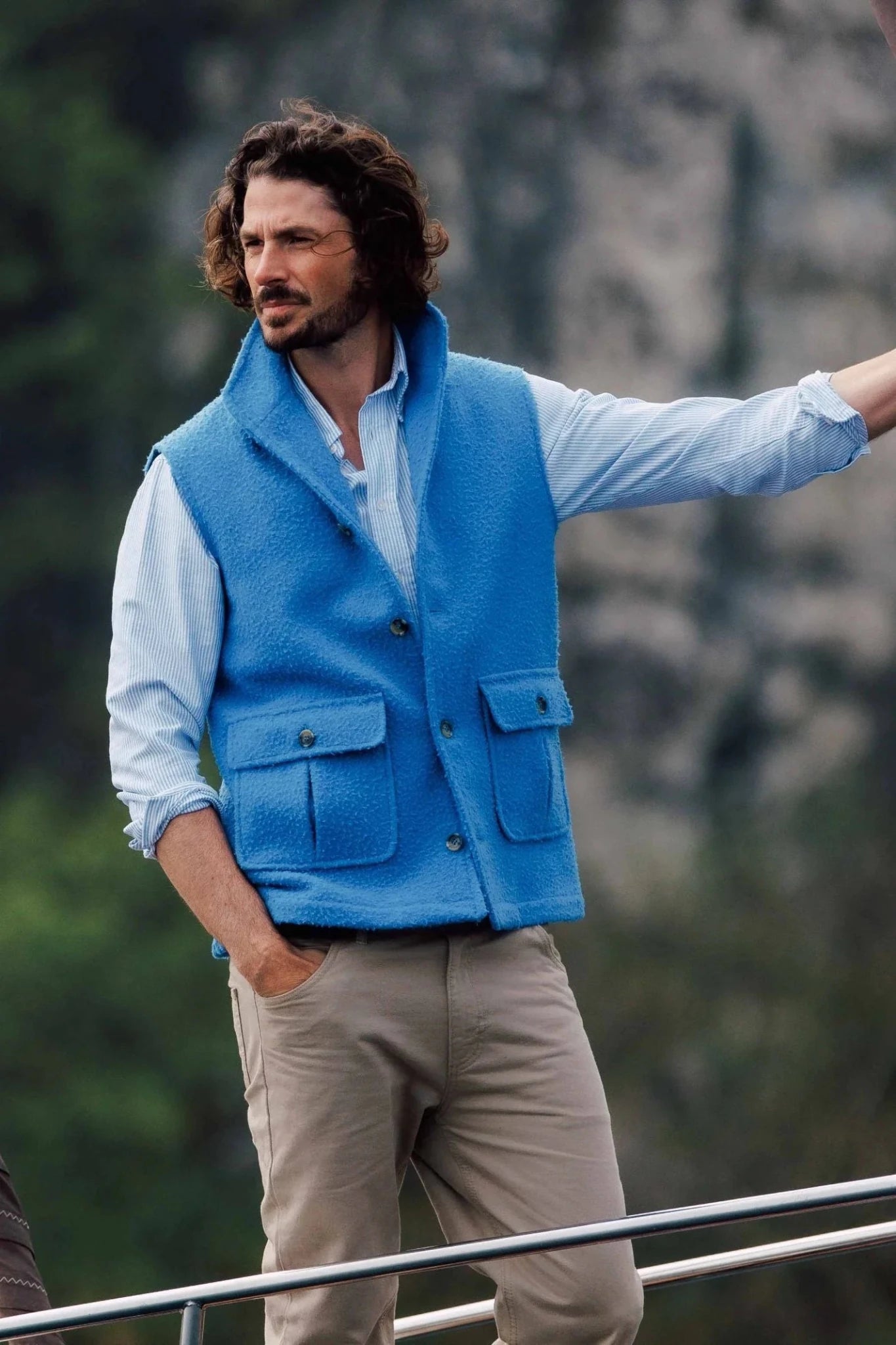A man with wavy brown hair and a beard stands outdoors in a light blue button-up, beige pants, and the Campbell's of Beauly Auld Stock Casentino Fleece Gilet in Blue Lovat. He holds a metal railing with greenery and rocks behind him.