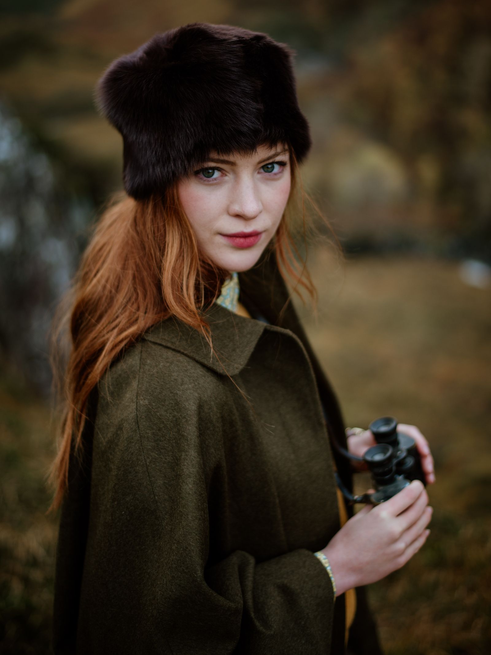 A woman with long red hair, wearing the Campbells of Beauly Loden Green Cape and a dark fur hat, stands outdoors holding binoculars against a blurred natural landscape.