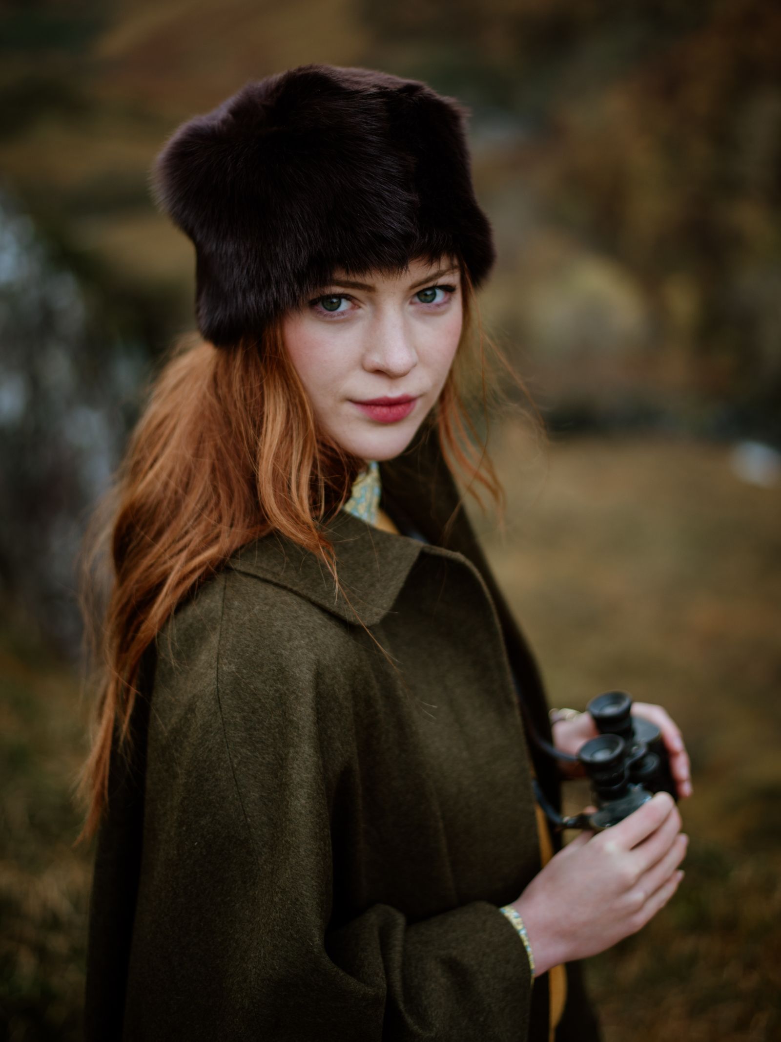 A woman with long red hair stands outdoors in natures autumn colors, wearing a Campbells of Beauly Pillar Box Hat and an olive green coat, holding binoculars.
