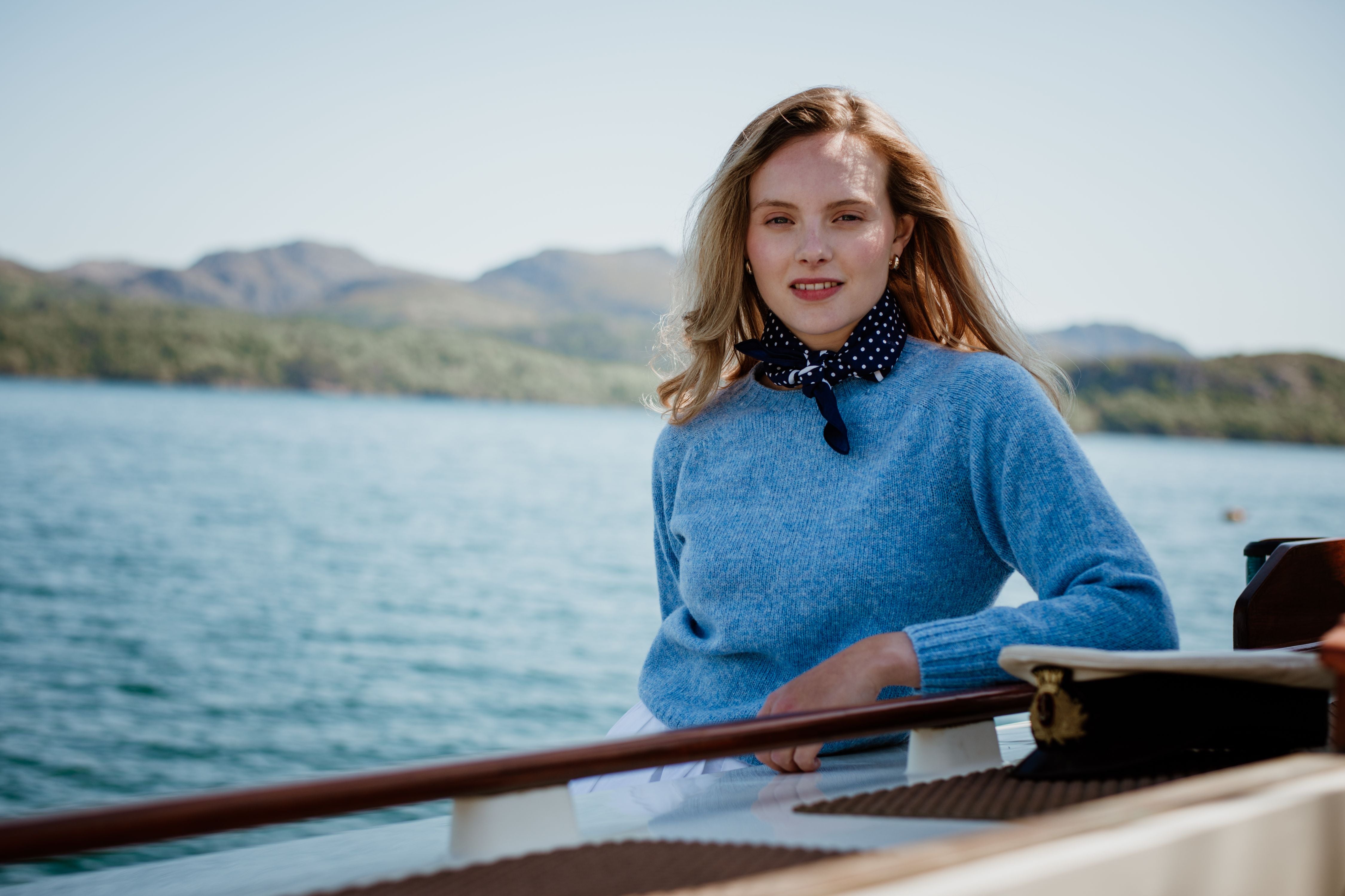 A woman wearing a blue sweater and a polka dot scarf leans on the railing of a boat, with calm water and distant green hills visible in the background under a clear sky.