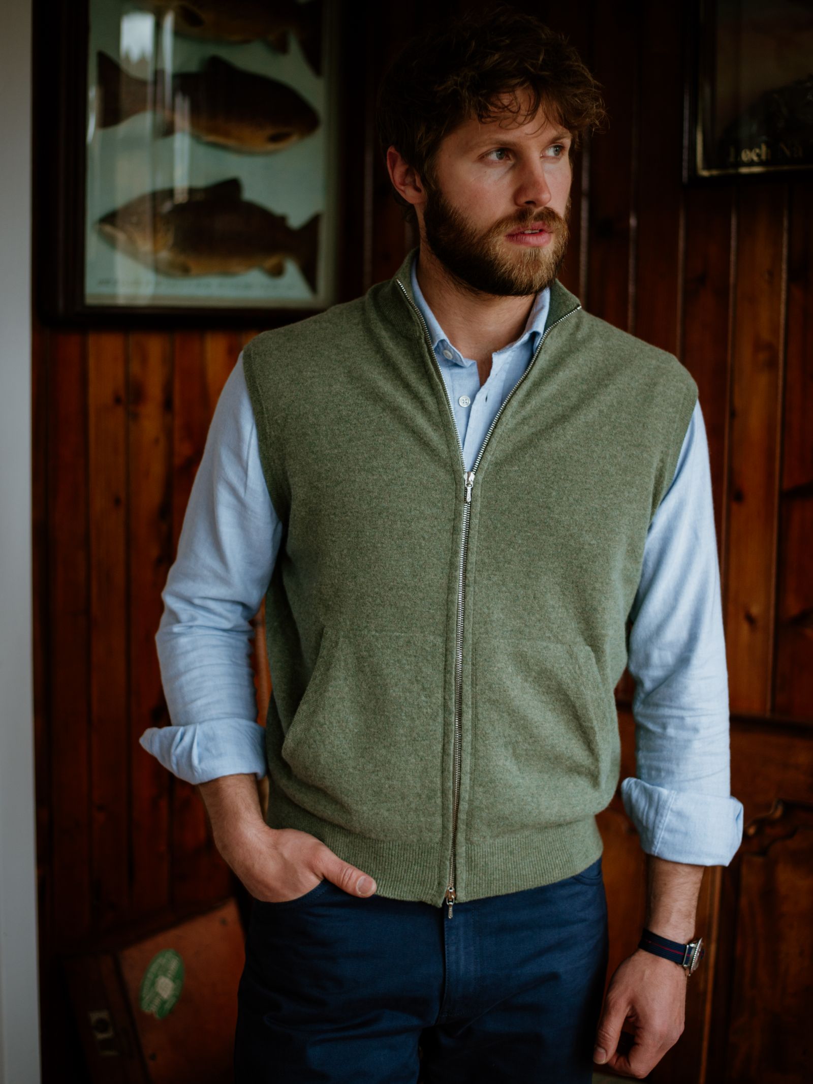 A man with a beard and tousled hair wears a Campbells of Beauly Zip Gilet in olive green over a light blue shirt, standing indoors against wooden walls adorned with framed fish illustrations, evoking timeless style.