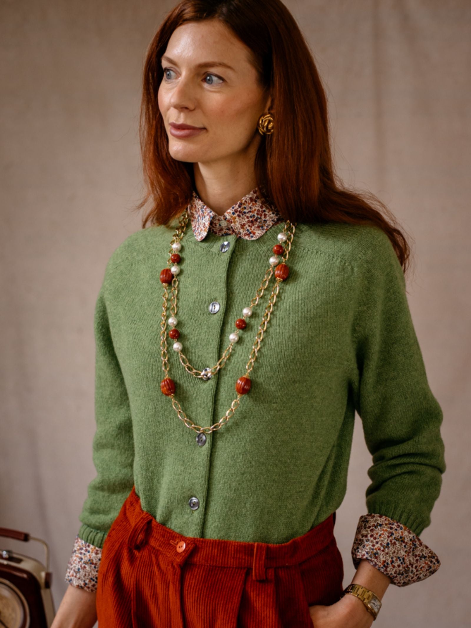 A woman with long auburn hair wears the Campbell’s of Beauly Crew Cardigan in green over a floral collar shirt, red corduroy pants, and layered gold jewelry, standing against a neutral background.