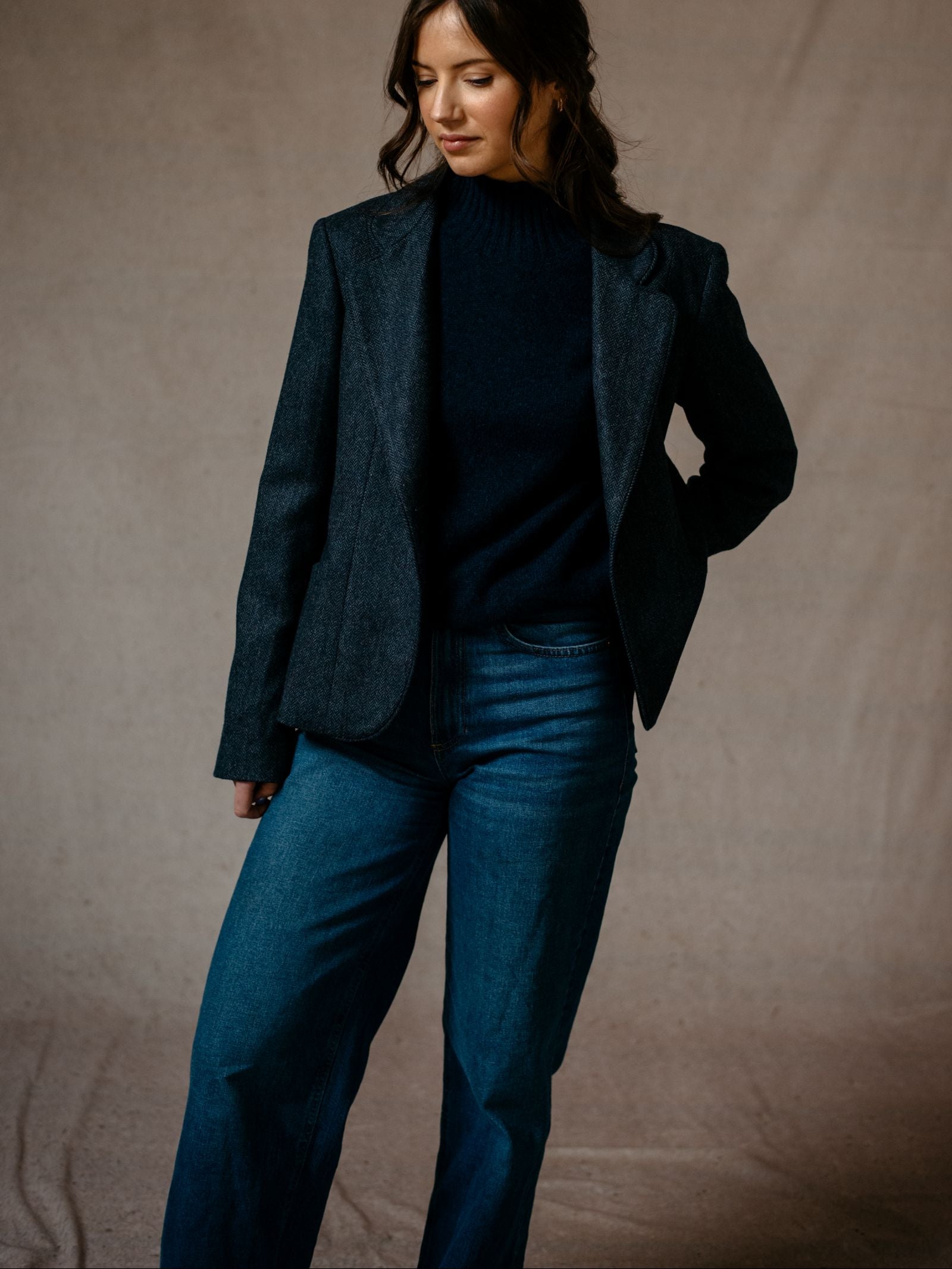 A woman with long, wavy brown hair stands in front of a neutral background wearing Campbells of Beauly’s House Jacket over a black sweater and wide-leg blue jeans, looking down with one hand in her pocket.