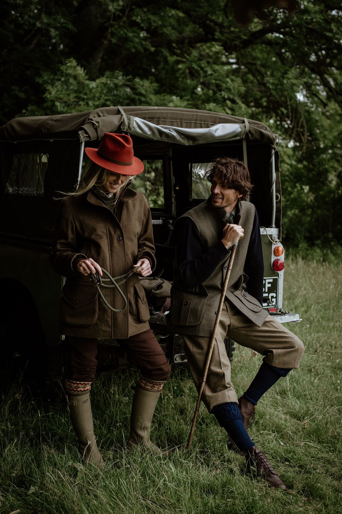 A man and a woman in country attire stand by a vintage Land Rover in a grassy field.