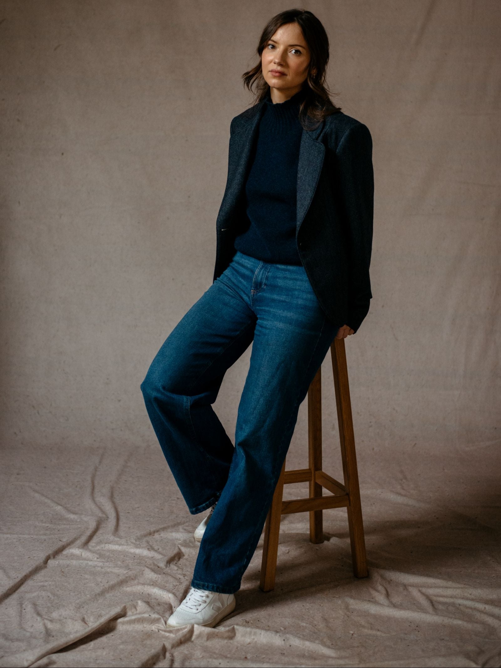 A woman with wavy dark hair sits on a wooden stool, wearing the House Jacket by Campbells of Beauly over a black turtleneck, blue jeans, and white sneakers, showcasing timeless style against a neutral fabric backdrop.