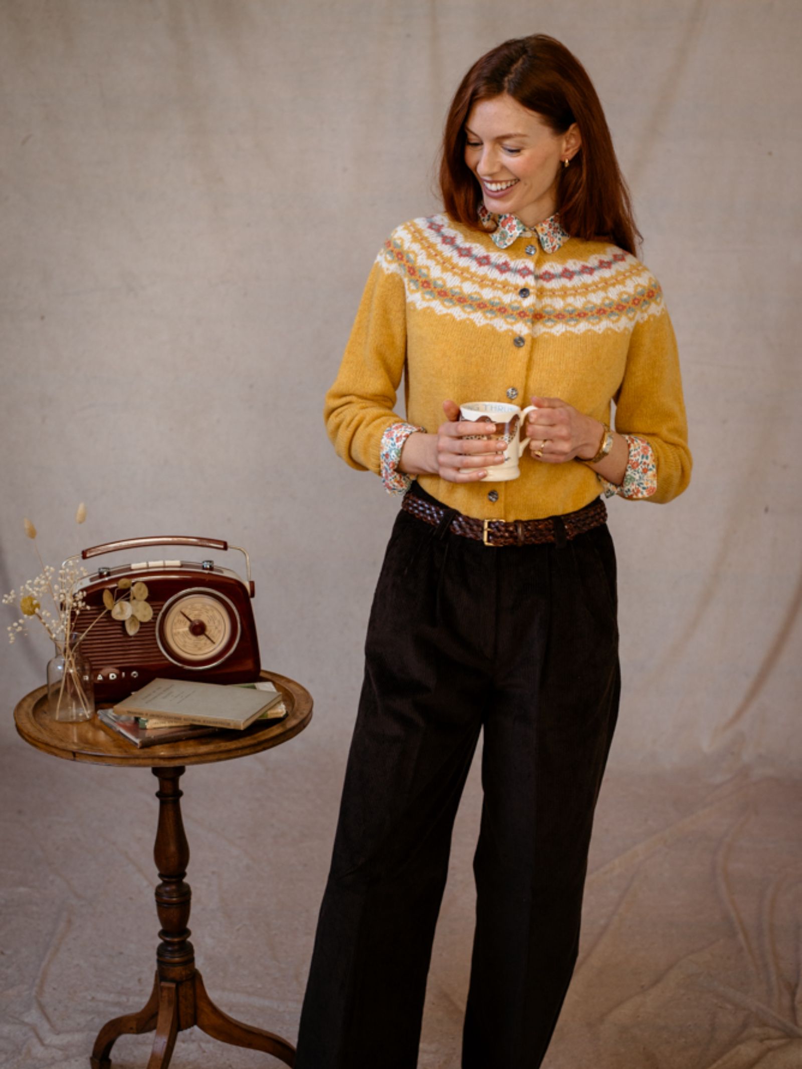 A woman wears the Campbells of Beauly Shetland Fairisle Cardigan in yellow with black pants, smiling as she holds a mug. Next to her, a small round table holds a vintage radio, dried flowers, and books against a neutral fabric backdrop.