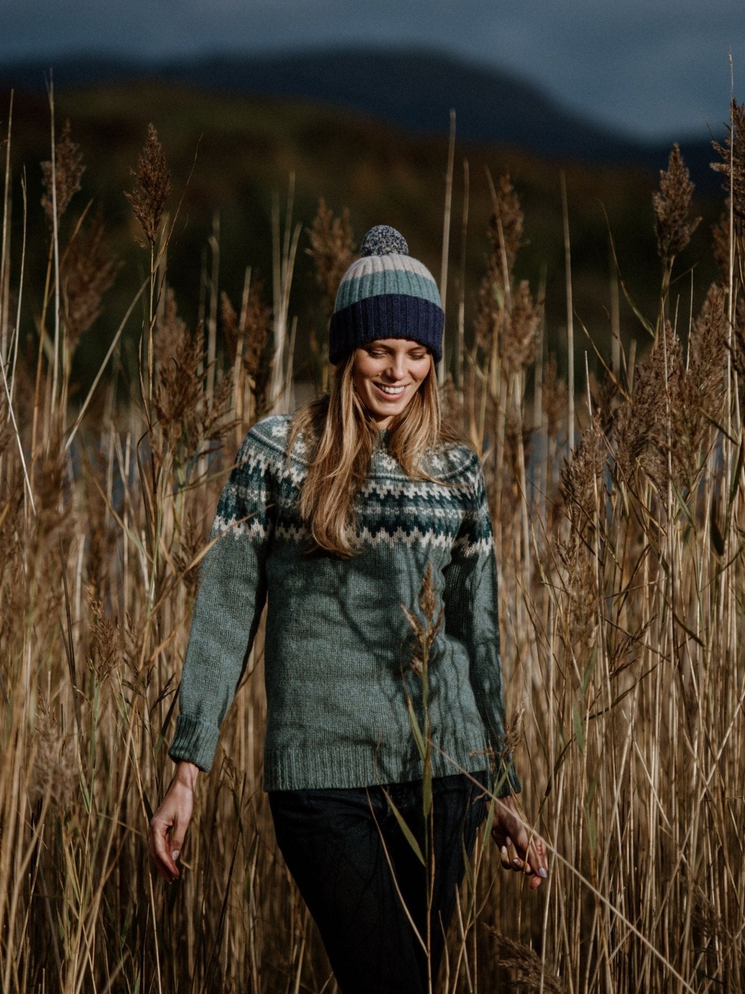 A woman in a green patterned sweater and Campbells of Beauly Lambswool Stripe Hat with Pom-Pom stands in tall golden grass, with rolling Scottish Borders hills and a dark sky behind her.