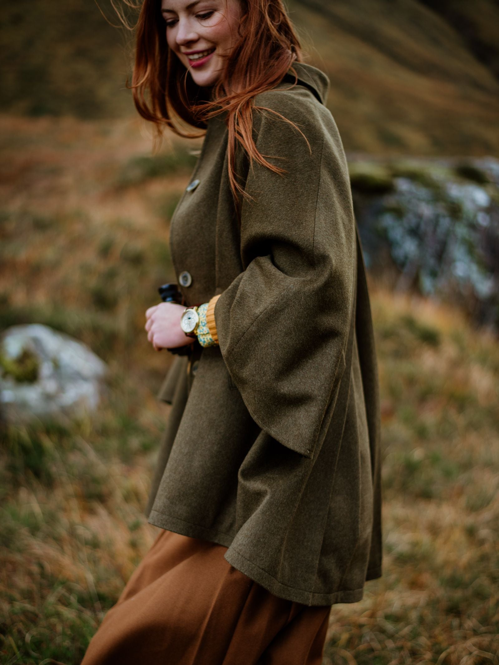 A woman with long red hair smiles outdoors on grassy terrain, wearing the Loden Green Cape by Campbells of Beauly and a brown skirt, holding binoculars in one hand.