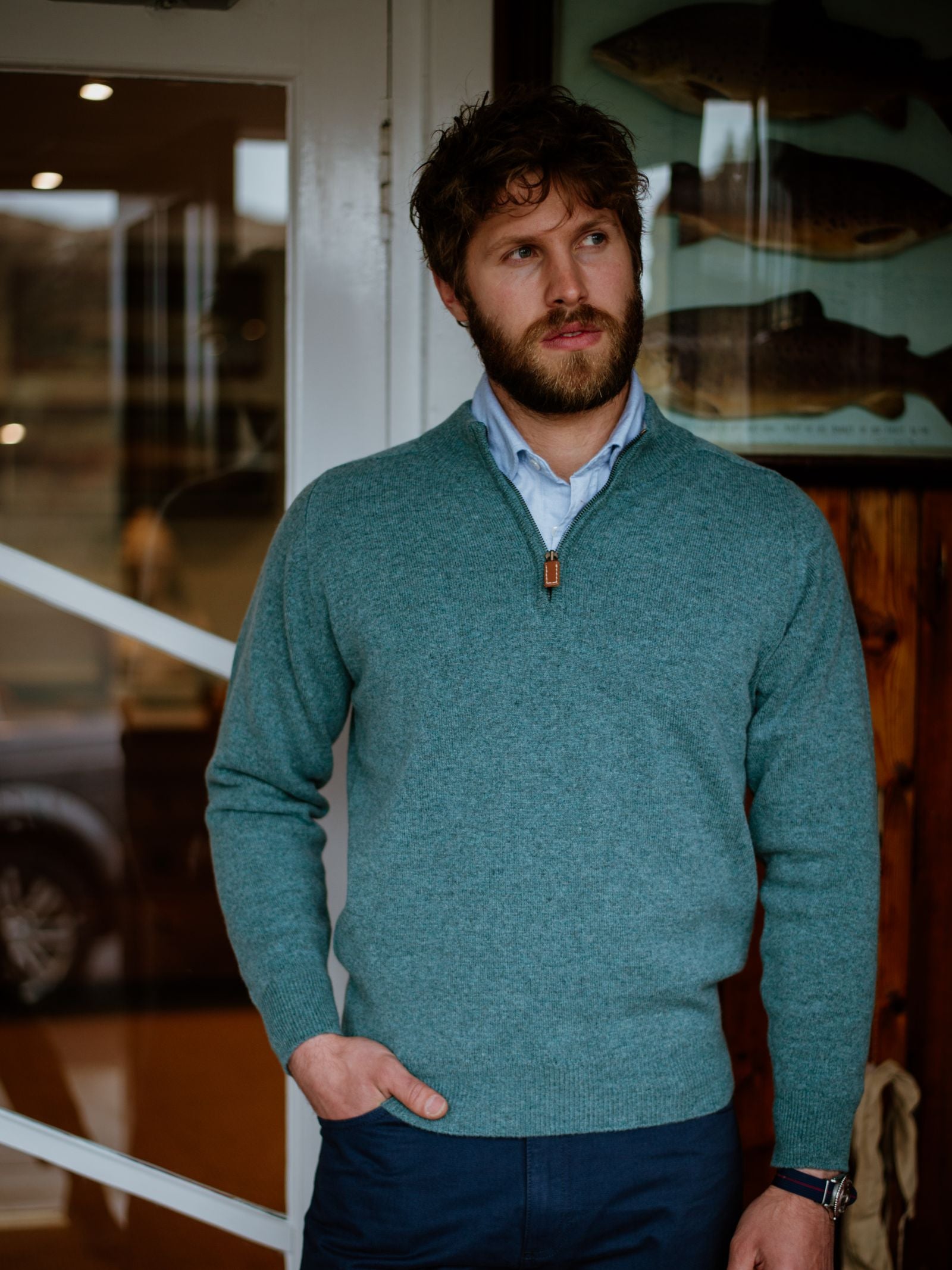 A man with curly brown hair and a beard stands indoors, wearing a teal Campbells of Beauly Lambswool Quarter Zip Jumper over a white shirt and navy pants. He looks to the side with one hand in his pocket; framed fish illustrations are visible behind him.