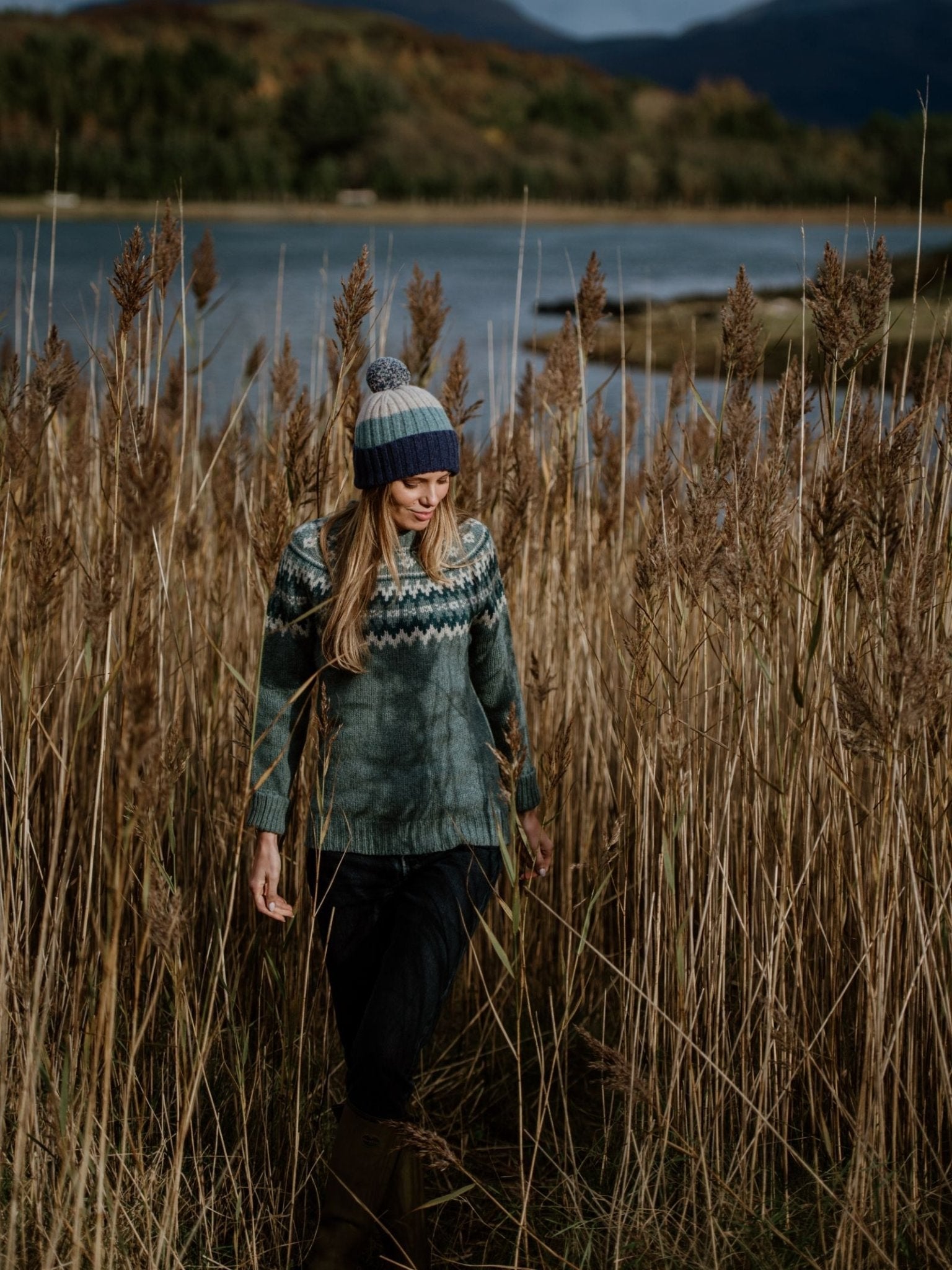 A woman in a patterned sweater and a Campbells of Beauly Lambswool Stripe Hat with Pom-Pom walks through tall reeds by a Scottish Borders lake, with trees and hills beneath a cloudy sky.