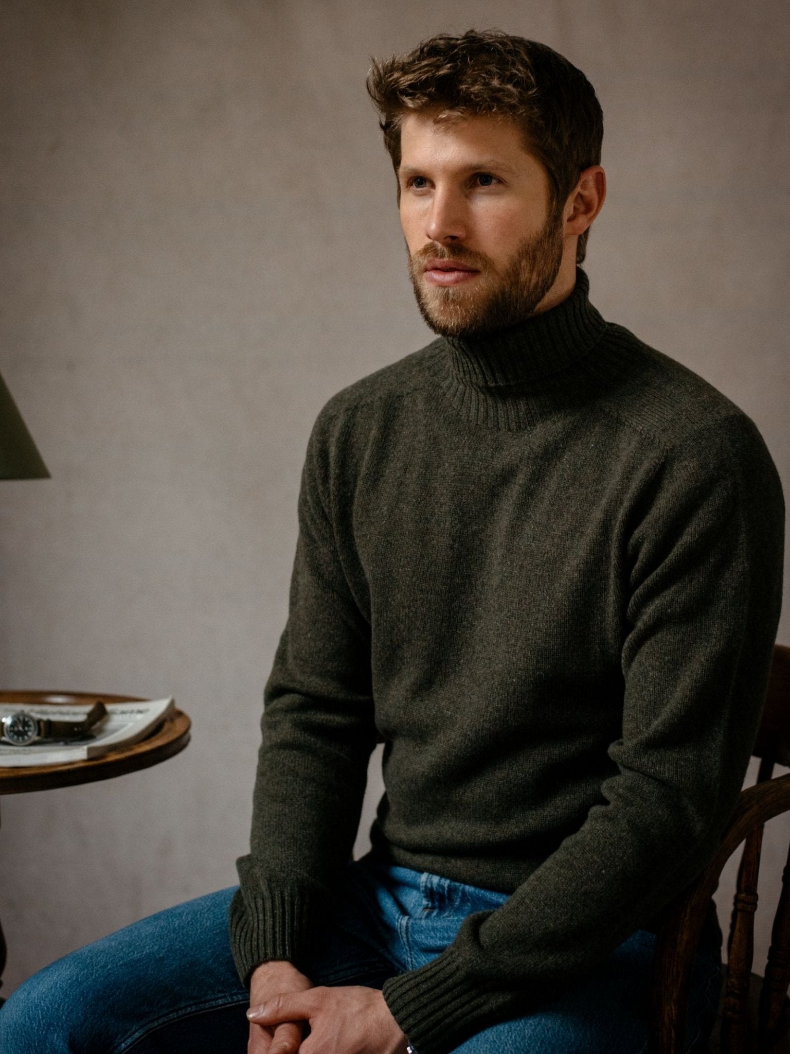 A man with short brown hair and a beard sits seriously on a wooden chair, wearing the Campbells of Beauly Lambswool Poloneck with blue jeans. A small table holding a lamp, watch, and papers is beside him.