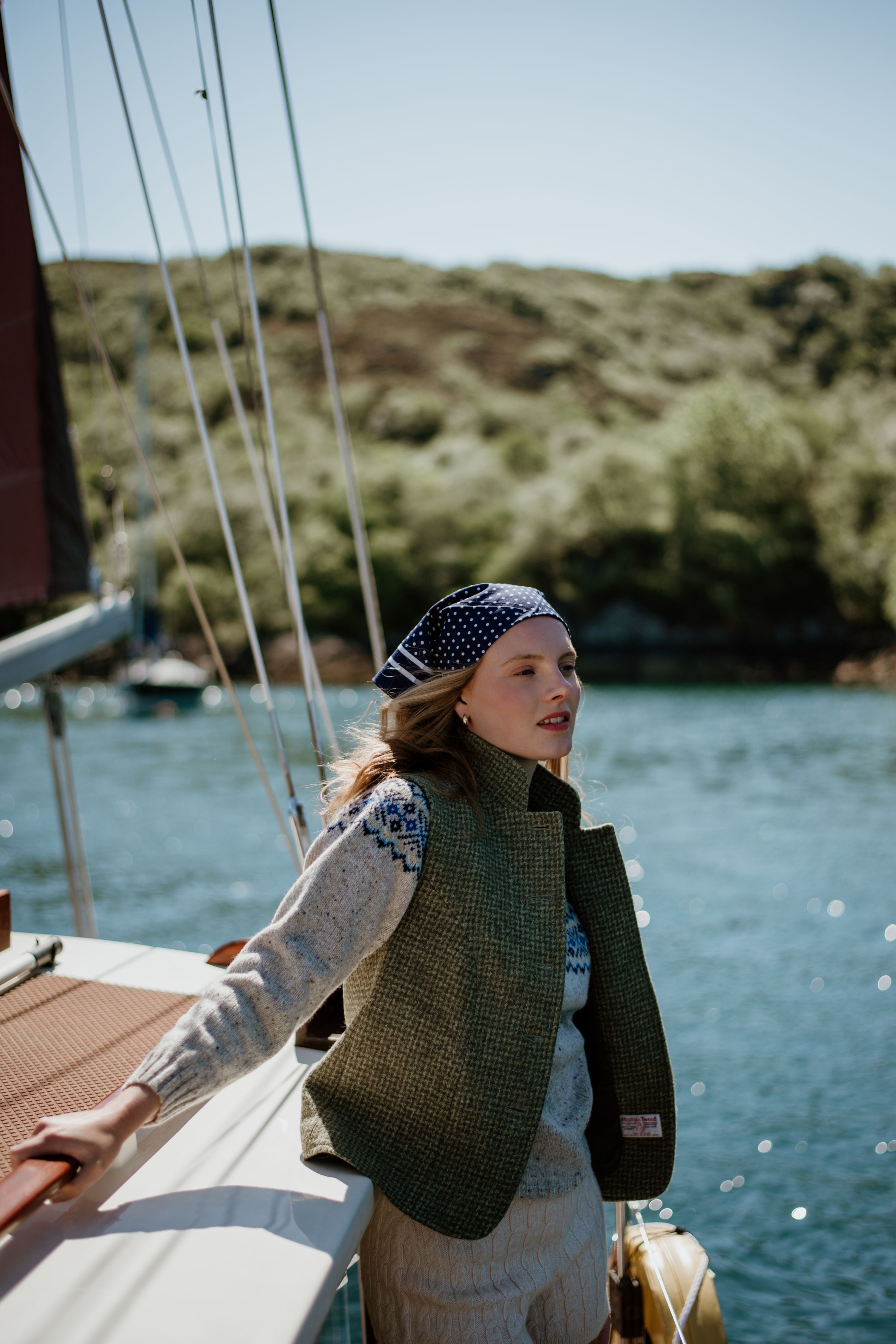 A woman wearing a blue patterned headscarf and green vest stands on a sailboat, holding the railing, with water and green hills in the background on a sunny day.