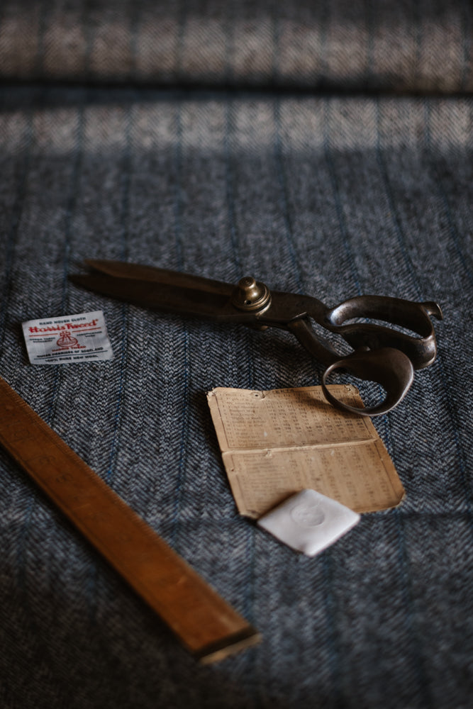 Large metal scissors, a wooden ruler, a small packet of needles, an old piece of folded paper, and a white square cloth rest on the textured grey herringbone House Tweed 95053 fabric by Campbell’s of Beauly.