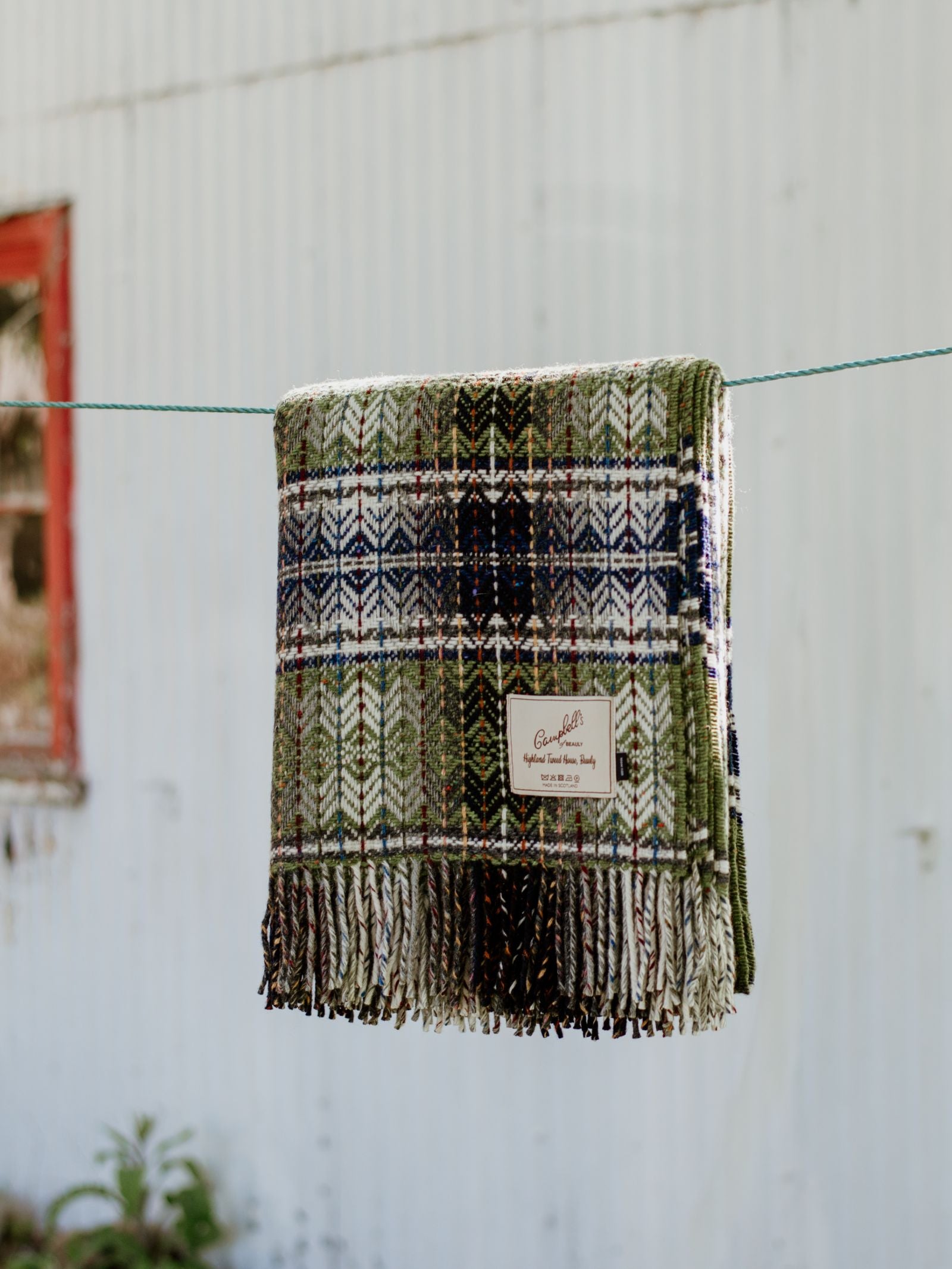 The Wyvis Lambswool Throw by Campbells of Beauly, featuring a green, blue, and cream plaid design with fringe in softly flecked Donegal lambswool, hangs outdoors against a white corrugated wall and red-trimmed window.
