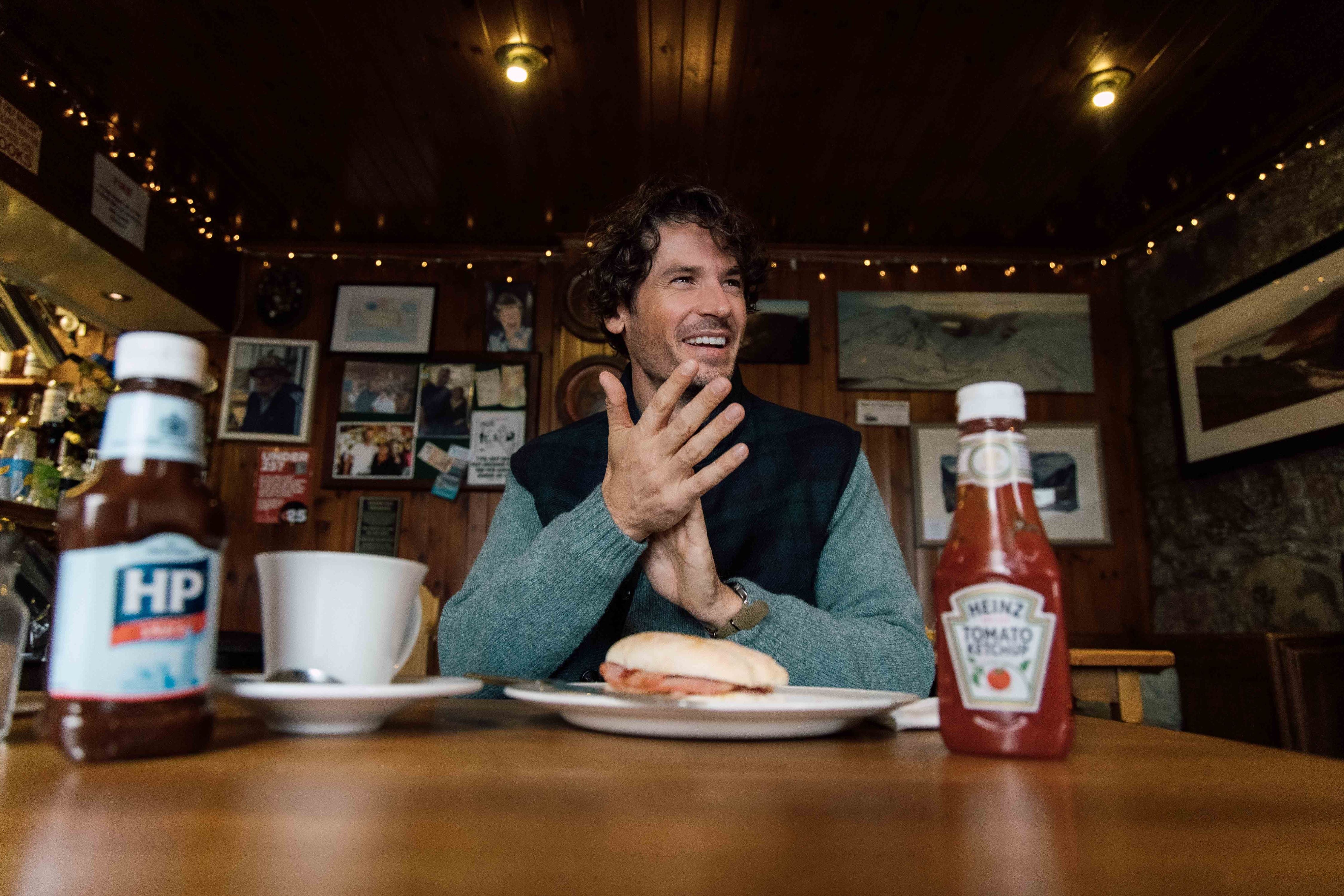 A man sits at a wooden table in a cozy cafe, smiling and clapping his hands. In front of him is a sandwich on a plate, a white mug, and bottles of HP Sauce and Heinz Tomato Ketchup. The background has photos and warm lighting.