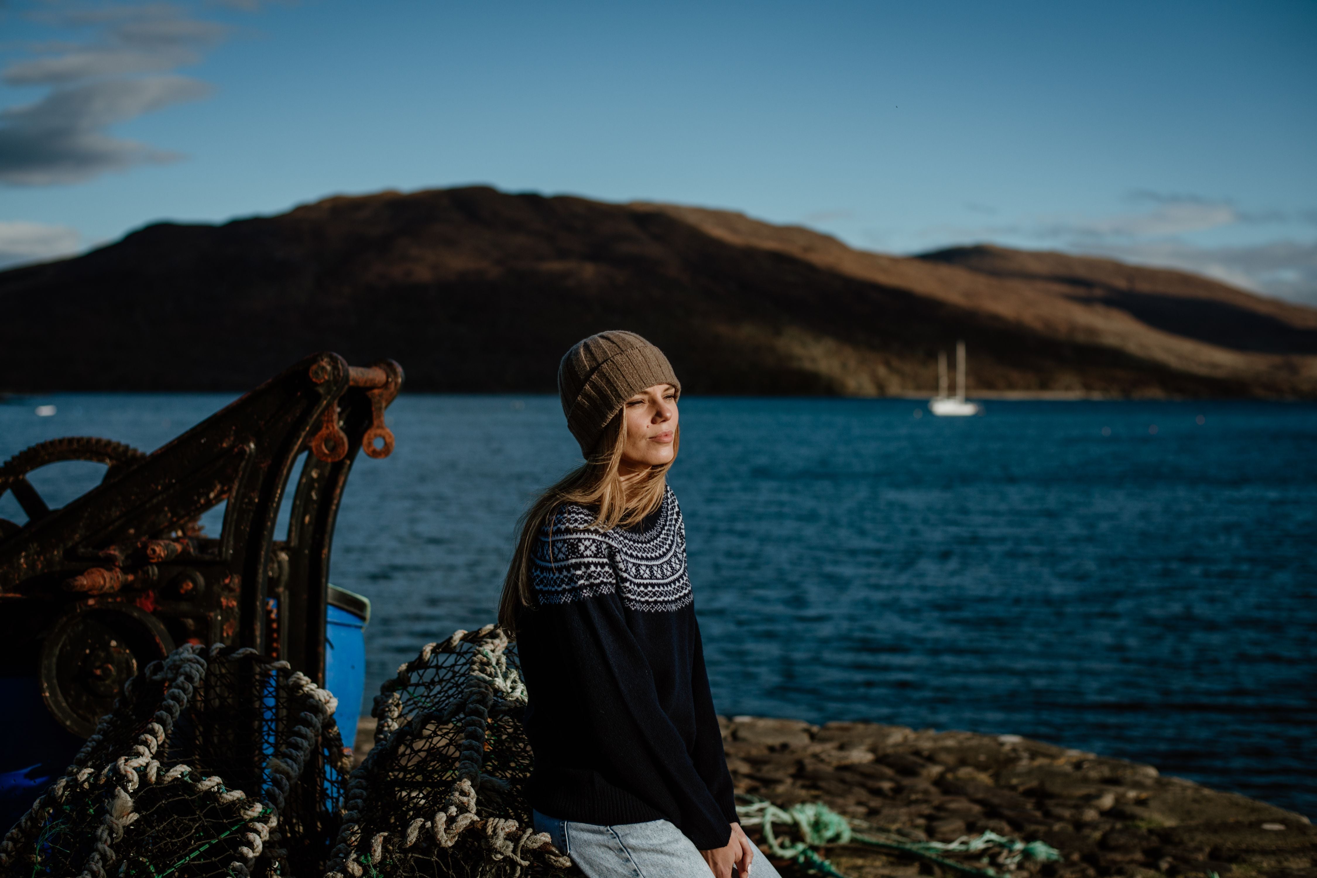 A woman in a knit hat and sweater sits by fishing nets on a rocky dock, with a calm blue lake and mountains in the background under a clear sky.
