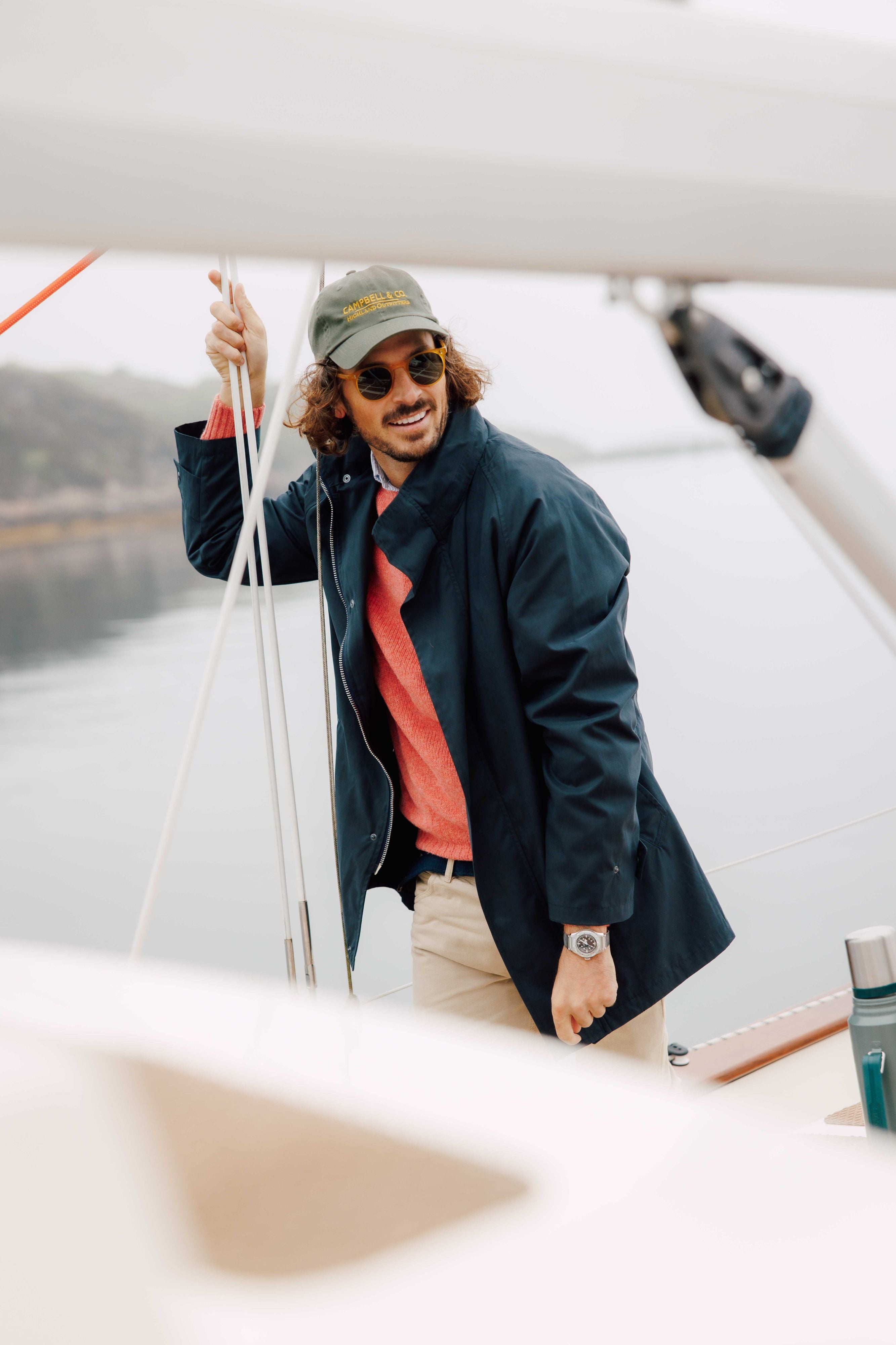 A man with curly hair, sunglasses, and a cap stands on a sailboat, smiling. He is wearing a navy coat over a red sweater and beige pants, with one hand holding a rope. The water and shoreline are visible in the background.