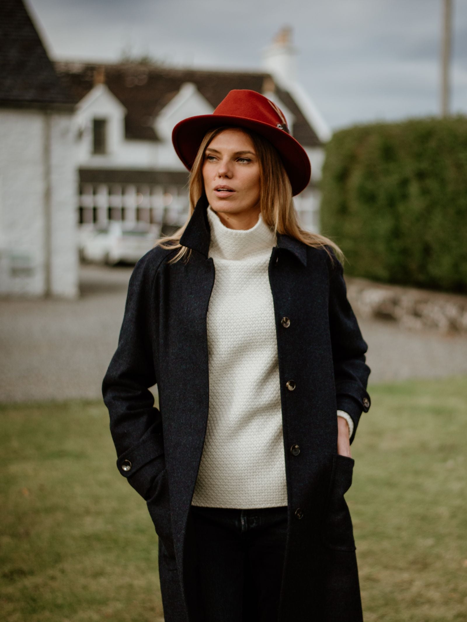A woman stands on grass with her hands in her pockets, wearing the Campbells of Beauly Midnight Collar Coat and a white sweater. Red hat tops off her look; white houses and greenery blur in the background.