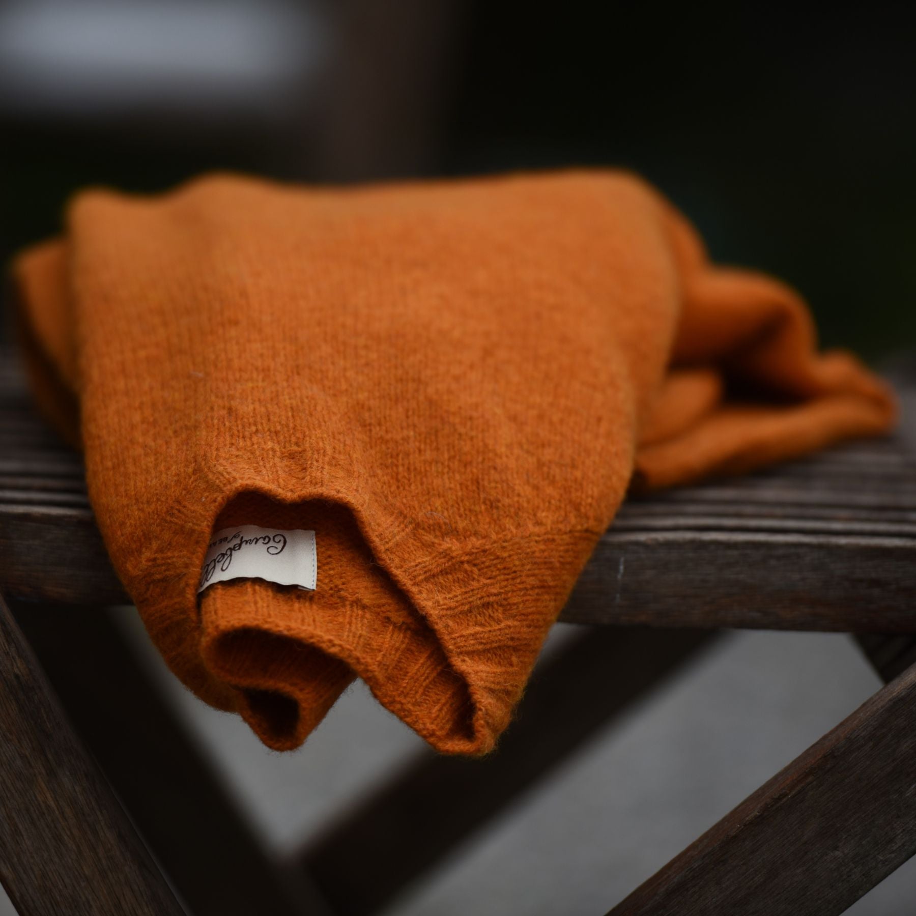 An orange knit sweater is folded and placed on a wooden surface. A small white tag with text is visible at the edge of the sweater. The background is blurred.