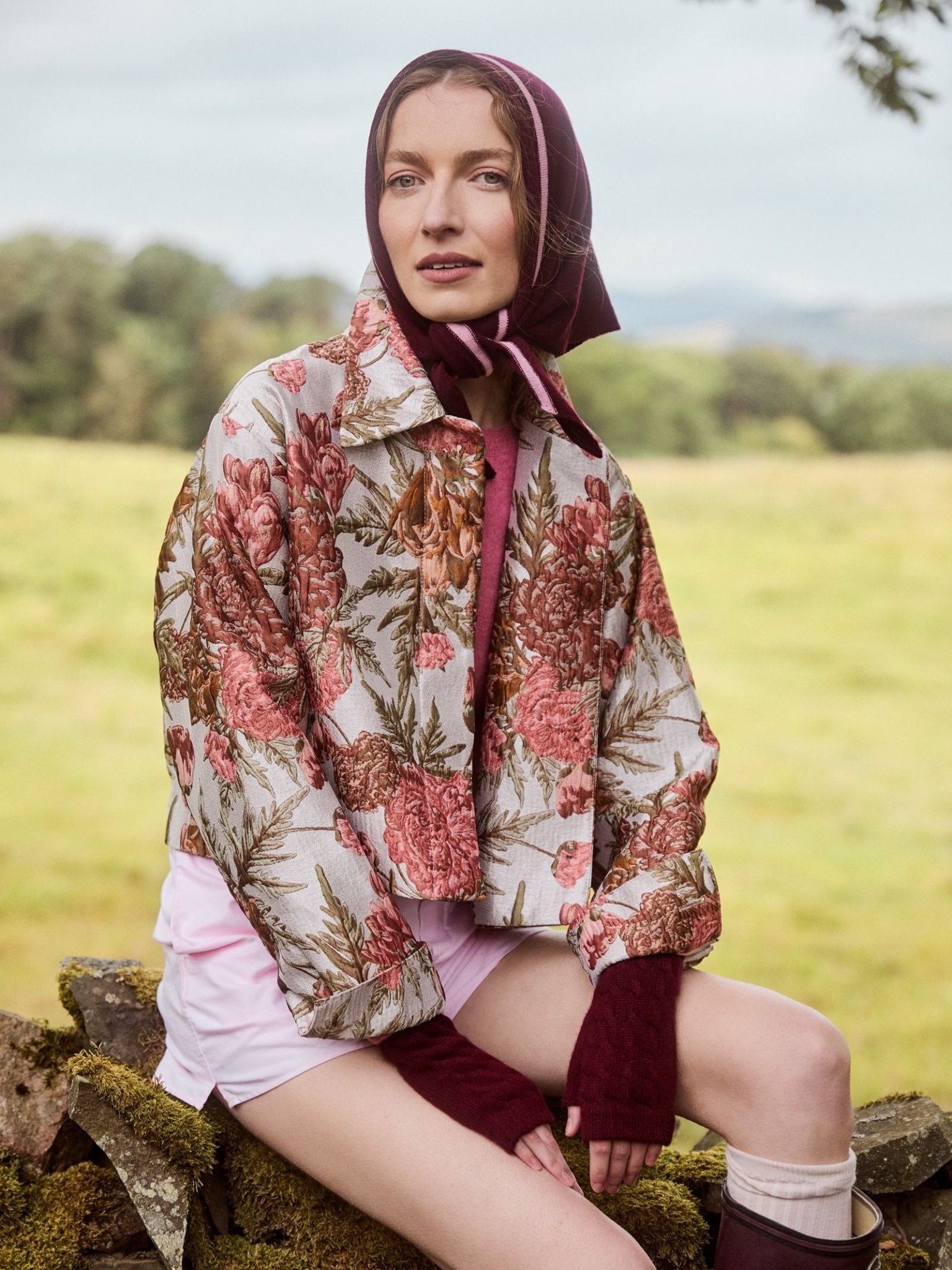 A woman wearing a floral jacket, pink shorts, fingerless gloves, and the Campbell’s of Beauly Cashmere Neckerchief in Damson sits on a stone wall in a grassy field, with trees and hills in the background.