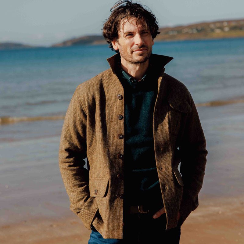A man with wavy hair stands on a sandy beach, wearing the Campbells of Beauly Casual Shacket over a dark sweater. The sea and distant coastline are visible in the background under a bright sky.