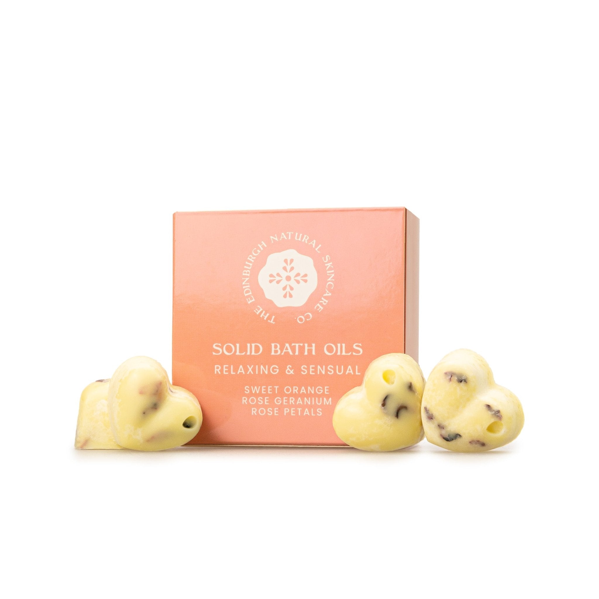 A box labeled Sweet Orange & Geranium Solid Bath Oils by The Edinburgh Natural Skincare Company sits behind three yellow, heart-shaped bath oils with rose petals, made from natural ingredients and essential oils.