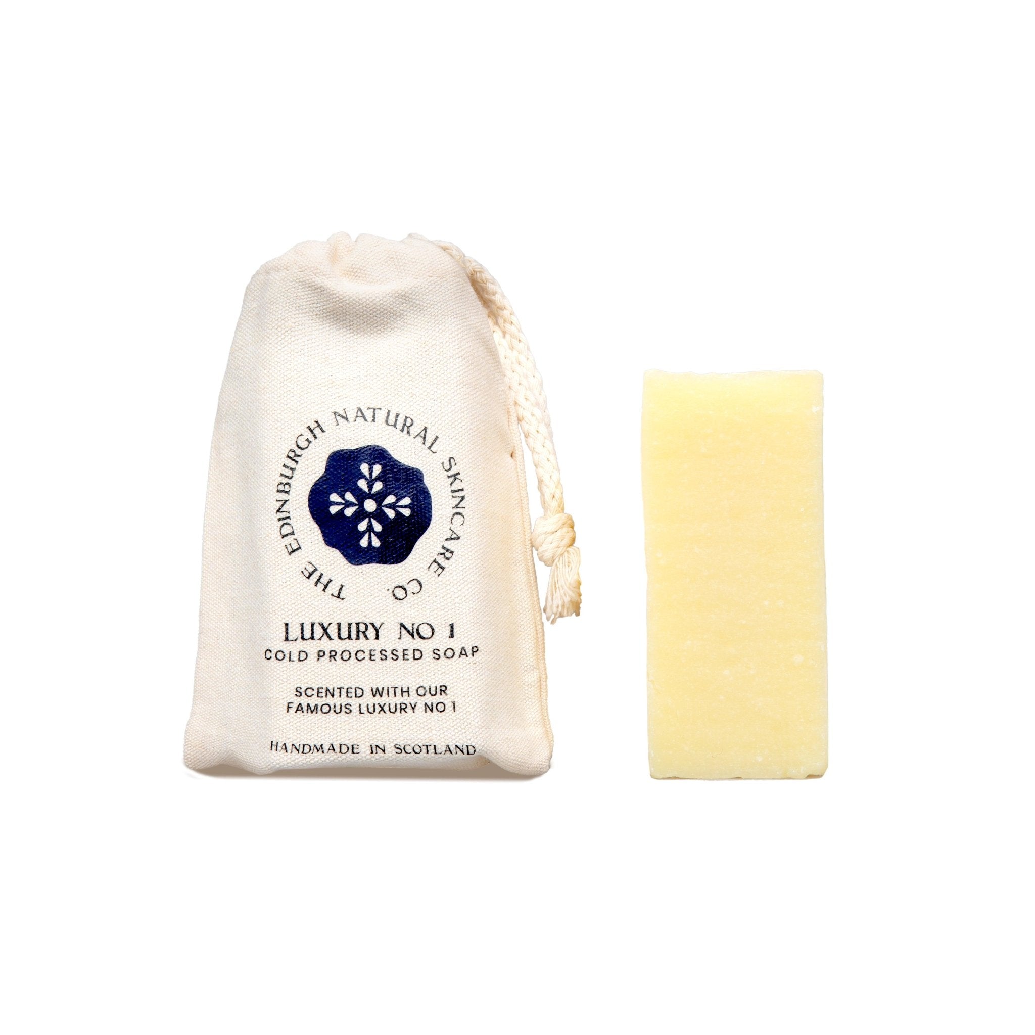 A rectangular yellow Luxury No 1. Face & Body Cleansing Soap by The Edinburgh Natural Skincare Company stands next to a cream drawstring bag featuring the brand’s blue floral logo.
