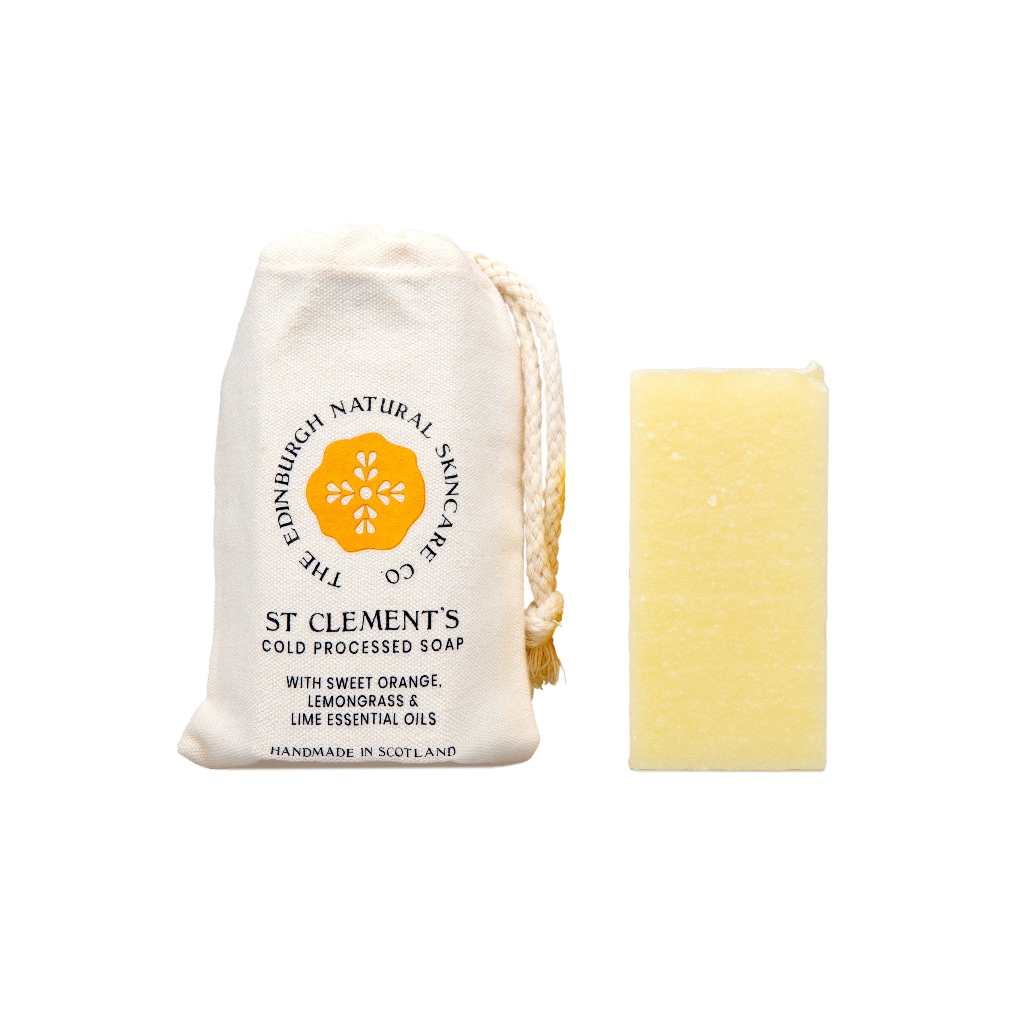 A yellow rectangular St Clement’s Cleansing Soap bar by The Edinburgh Natural Skincare Company stands upright beside a cream drawstring pouch labeled with an orange lemon slice, “100% natural oils,” and “Handcrafted in Scotland.”.