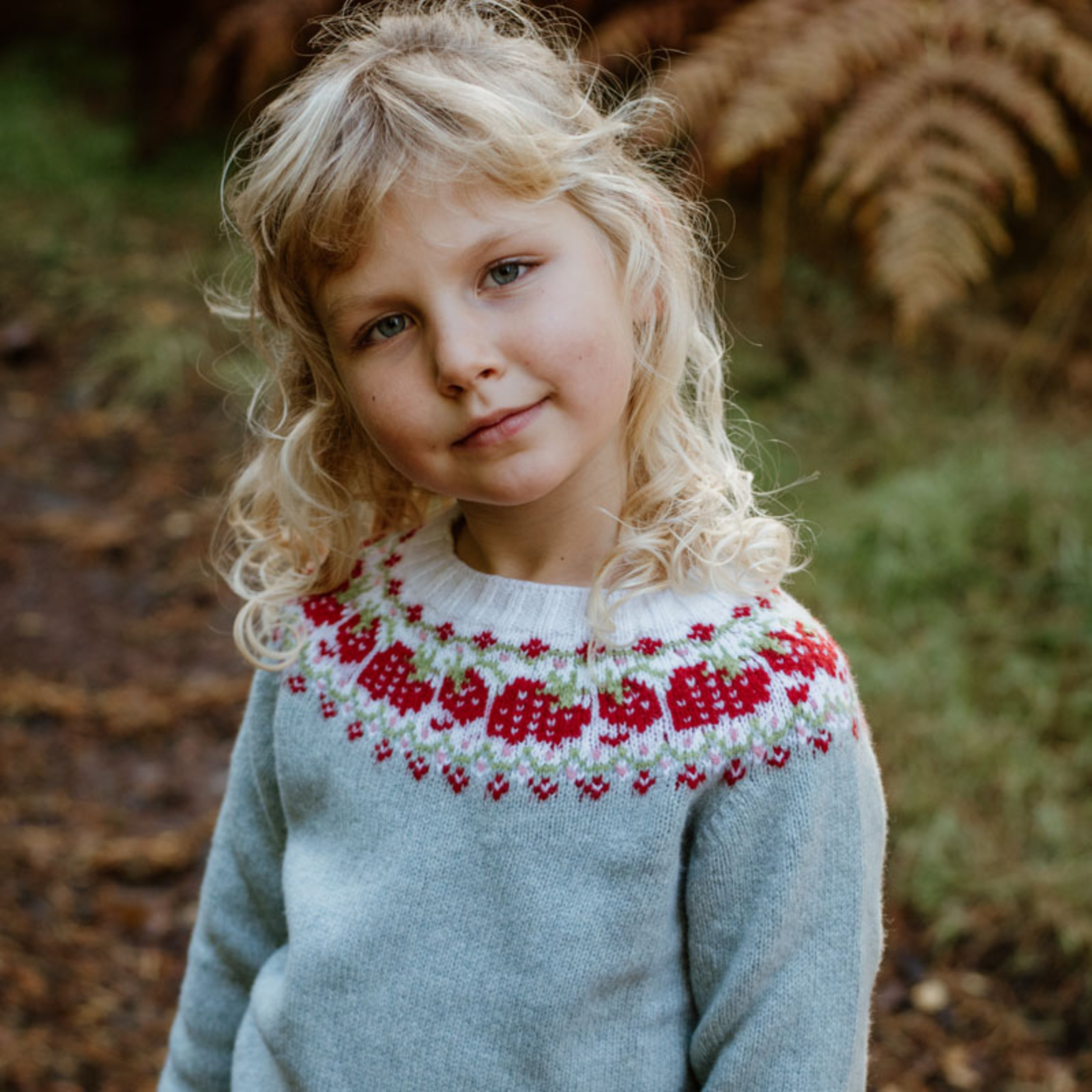 Young child with curly blonde hair wearing a light blue Campbell’s of Beauly sweater with a red and white patterned yoke, standing outdoors on a path with autumnal fern leaves in the background.