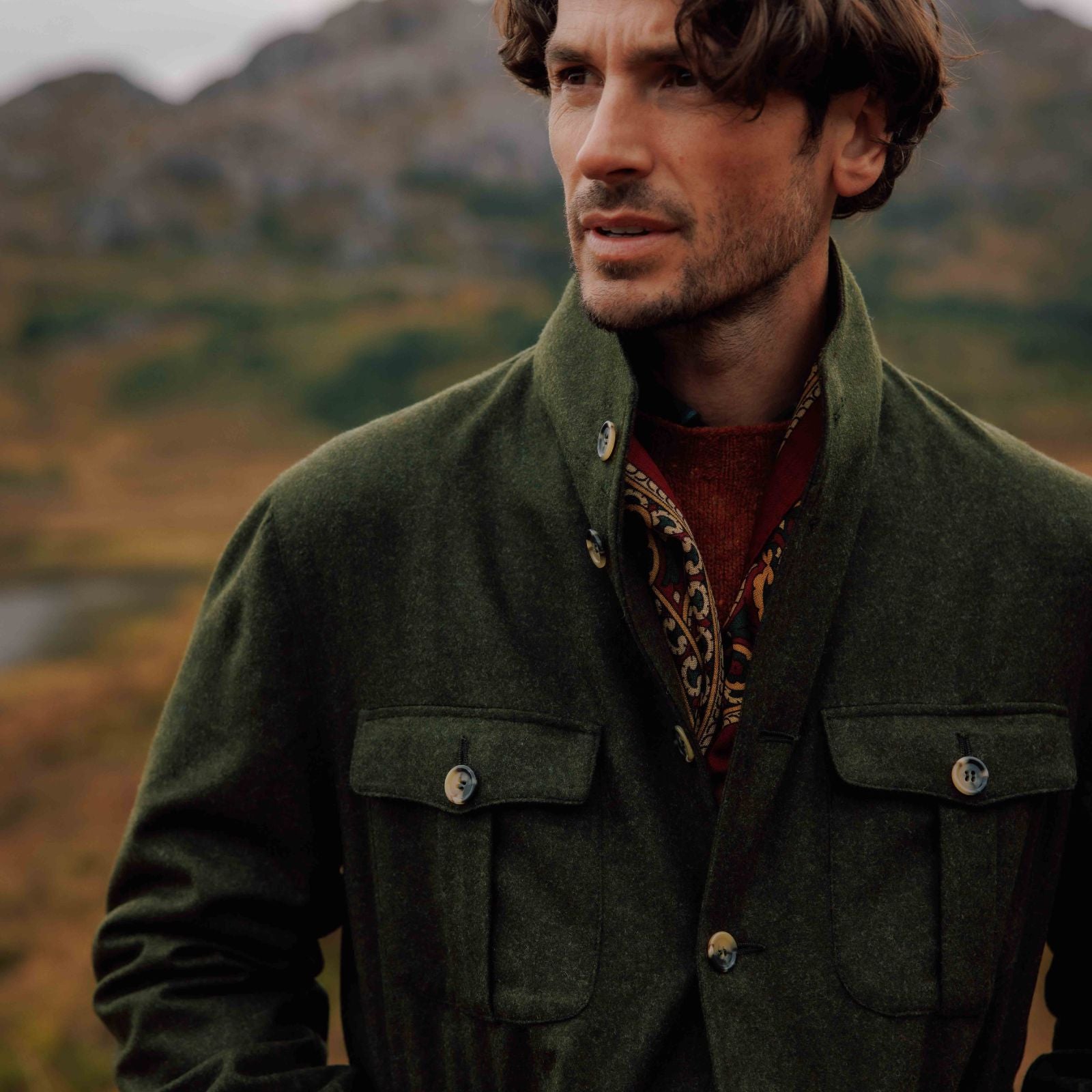 A man with wavy brown hair wearing a green jacket and patterned scarf from Campbells of Beauly stands outdoors, with blurred hills and greenery in the background.