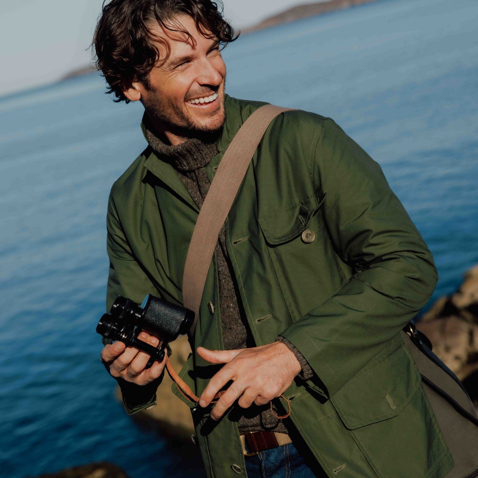 A man wearing a green jacket and brown shoulder strap from Campbells of Beauly smiles while holding a camera, standing outdoors near a body of water with rocky shoreline visible in the background.