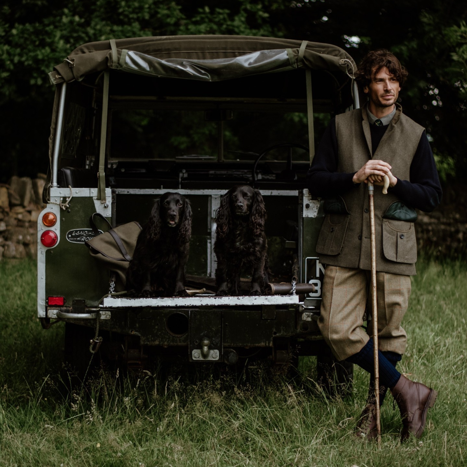 A man in country clothing leans on a walking stick beside an open Land Rover, with two black spaniels sitting on the vehicle’s tailgate in a grassy field.