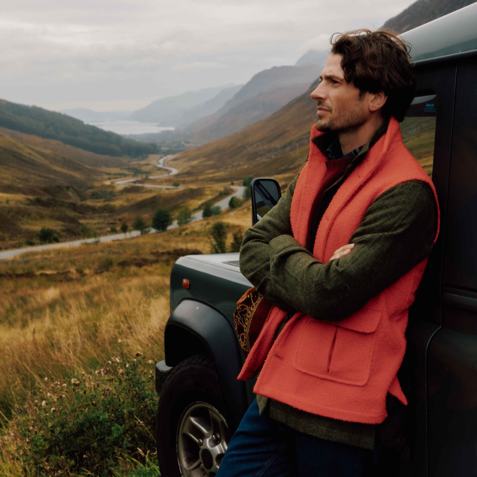 A man in a red vest and green sweater leans against a dark SUV, arms crossed, looking thoughtfully at a winding road through a scenic, hilly landscape under a cloudy sky.