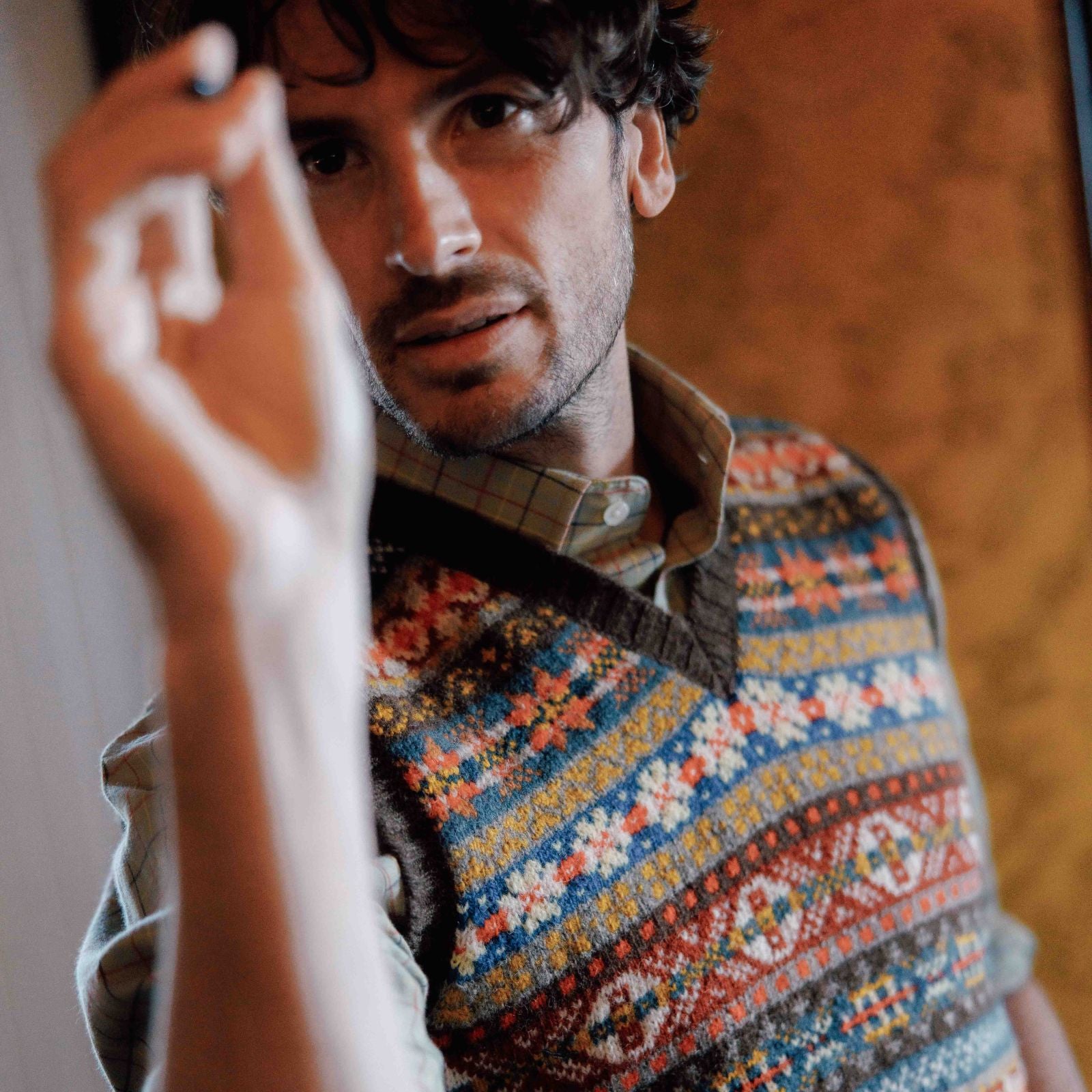 A man with wavy hair and stubble wears a colorful Campbells of Beauly patterned sweater vest over a plaid shirt. He faces the camera, holding up a marker or pen, as if writing or drawing on a surface in front of him.