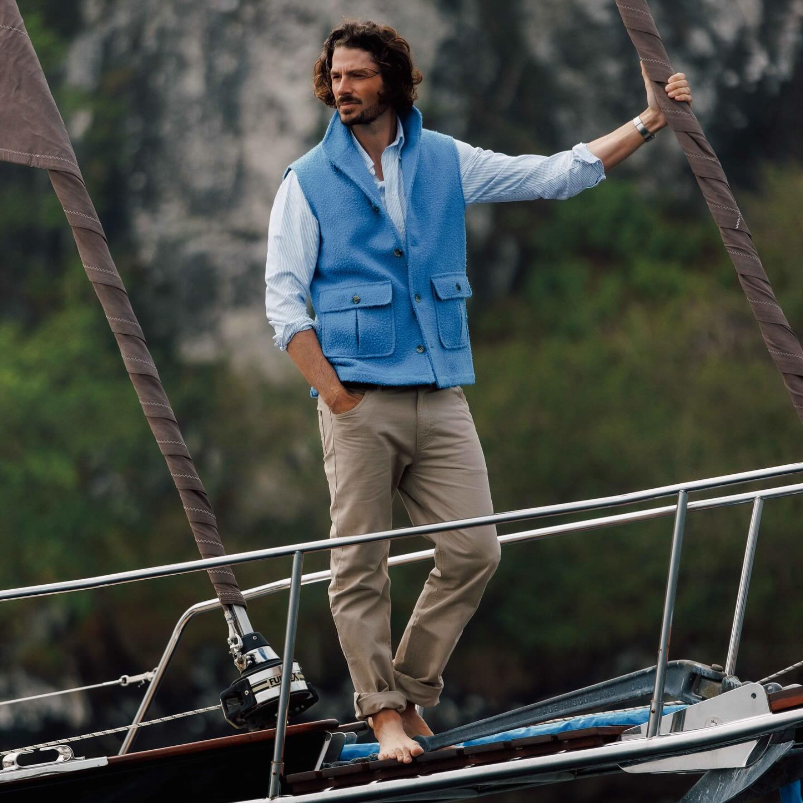 A barefoot man with curly hair, wearing a light blue vest and beige pants, stands on the deck of a sailboat, looking into the distance with one hand on the mast. Lush greenery and rocky cliffs are visible in the background.