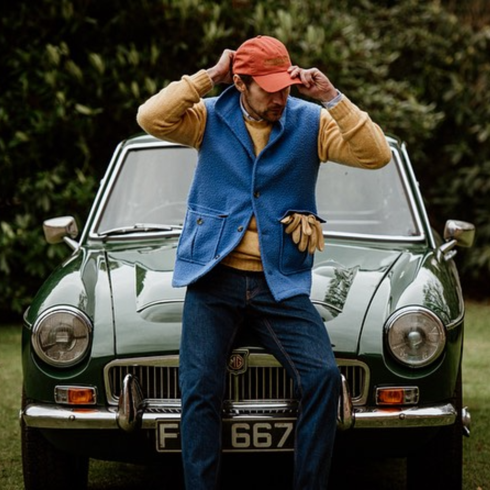 A man in a red cap, blue vest, yellow sweater, and jeans sits on the hood of a classic green MG car, adjusting his hat with both hands. Yellow gloves hang from his vest pocket. Lush greenery is in the background.