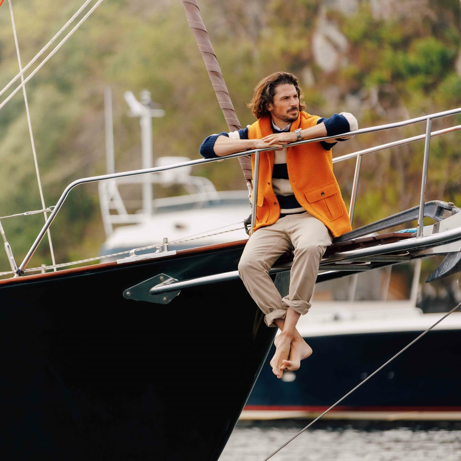 A man in an orange Campbells of Beauly vest sits barefoot on the bow of a sailboat, leaning on the railing and gazing thoughtfully into the distance, with water and blurred boats in the background.