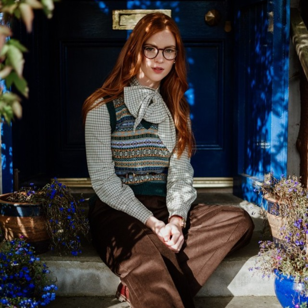 A young woman with long red hair and glasses sits on stone steps outside a blue door, wearing a patterned vest, checked blouse with a bow, and brown pants. Two baskets with purple flowers are beside her.